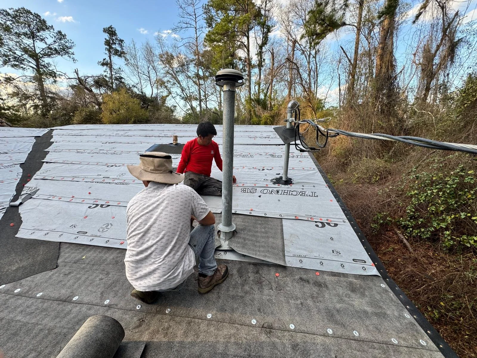 Two men installing a protective reflective roofing material on a roof surrounded by trees.