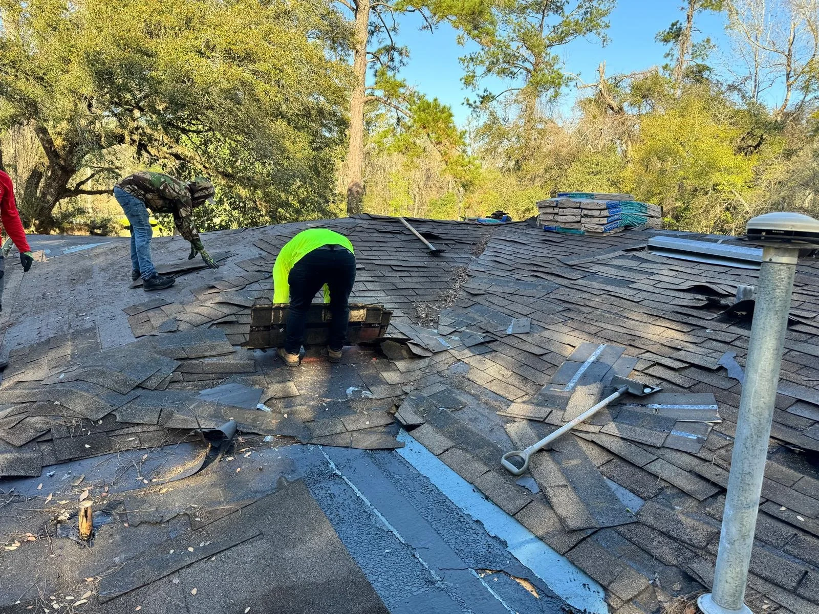 Workers repairing a damaged shingle roof, some shingles are removed and stacked on the roof, tools are scattered across the roof, and trees are visible in the background.
