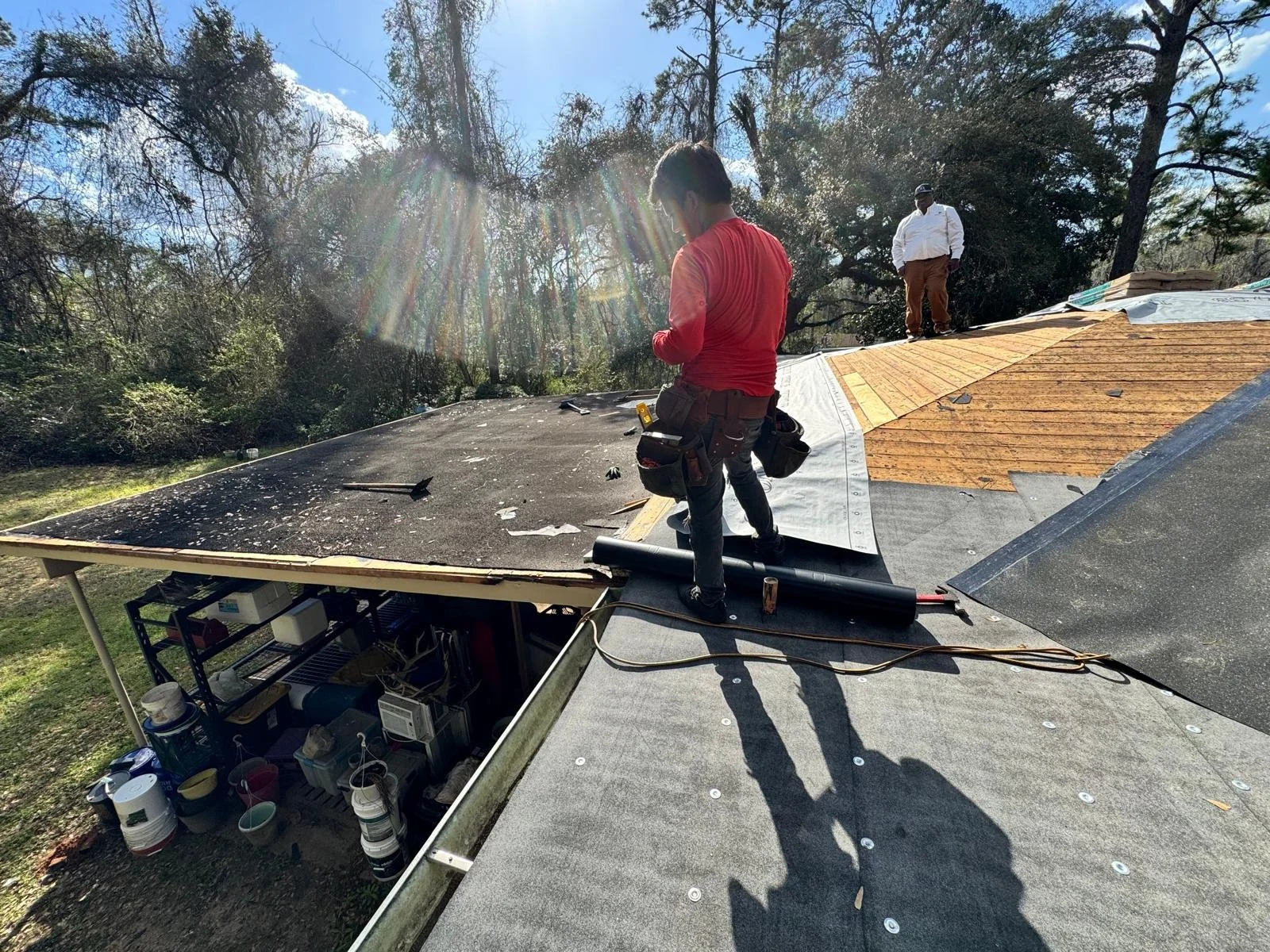 Two construction workers are installing roofing materials on a house with a forested backdrop and clear sky. One worker is standing near the edge of the roof, while the other is further back on the roof, handling a roll of roofing material.