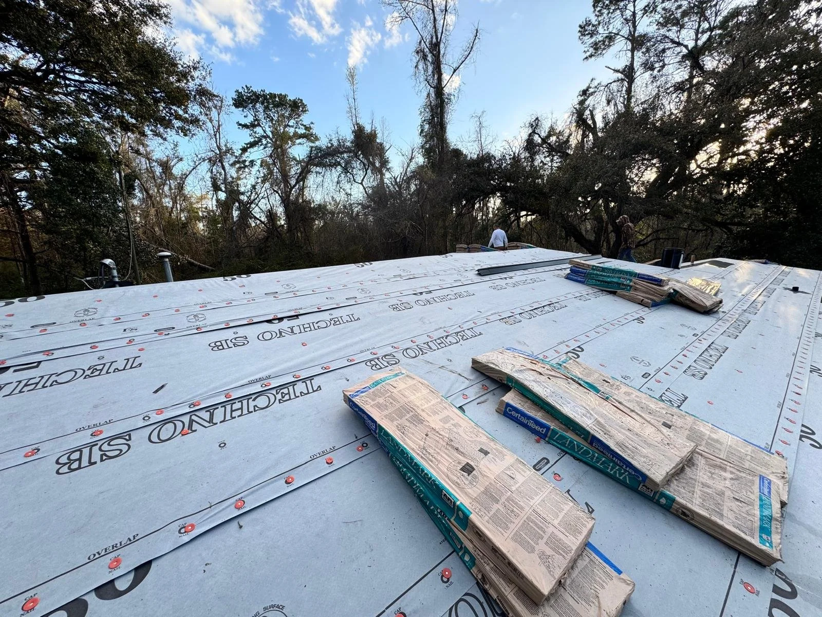 Roof under construction with Tietech roofing membrane and bundles of shingles, set against a background of trees and a partly cloudy sky.