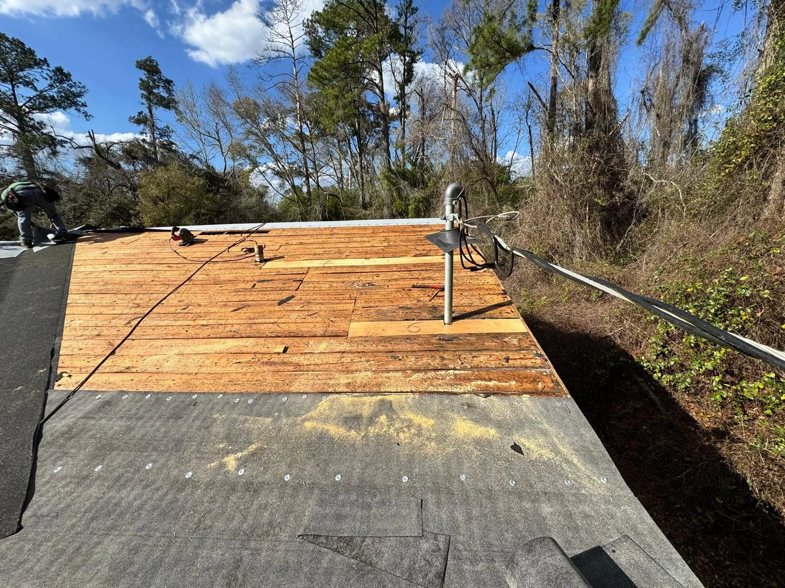 A construction worker installing or repairing a roof with uncovered wooden planks, a green safety harness, and wiring, on a partly cloudy day in a wooded area.
