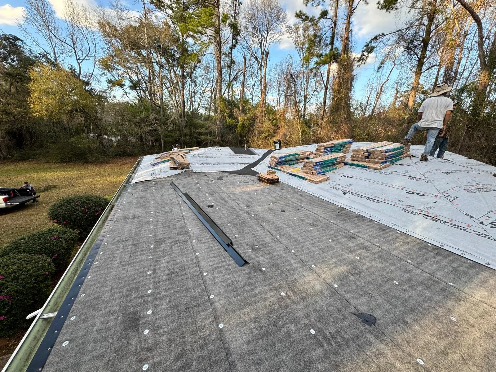 Construction workers on a roof with roofing materials and tools, surrounded by trees and a grassy yard with bushes and a truck.