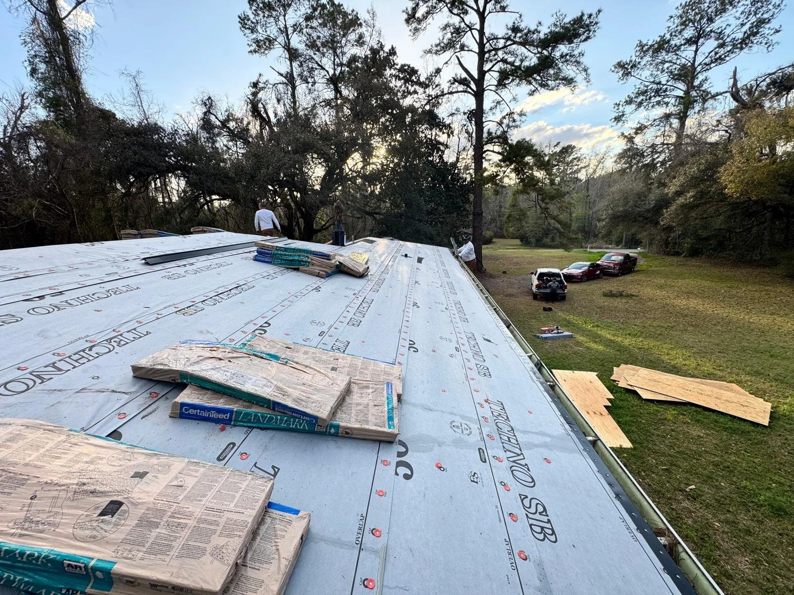 Roof under construction with roofing materials and tools, two workers, cars parked on grassy area, trees and sky in background.