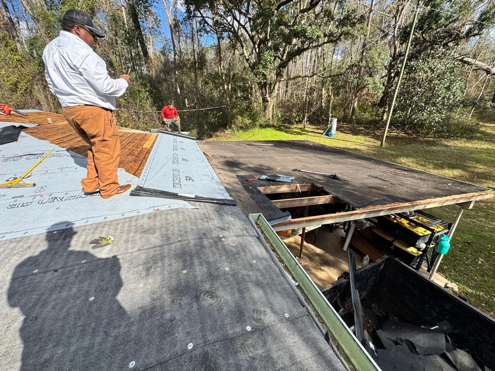 Workers installing roofing shingles on a house, with one worker standing on the roof and another below, surrounded by trees and construction tools.