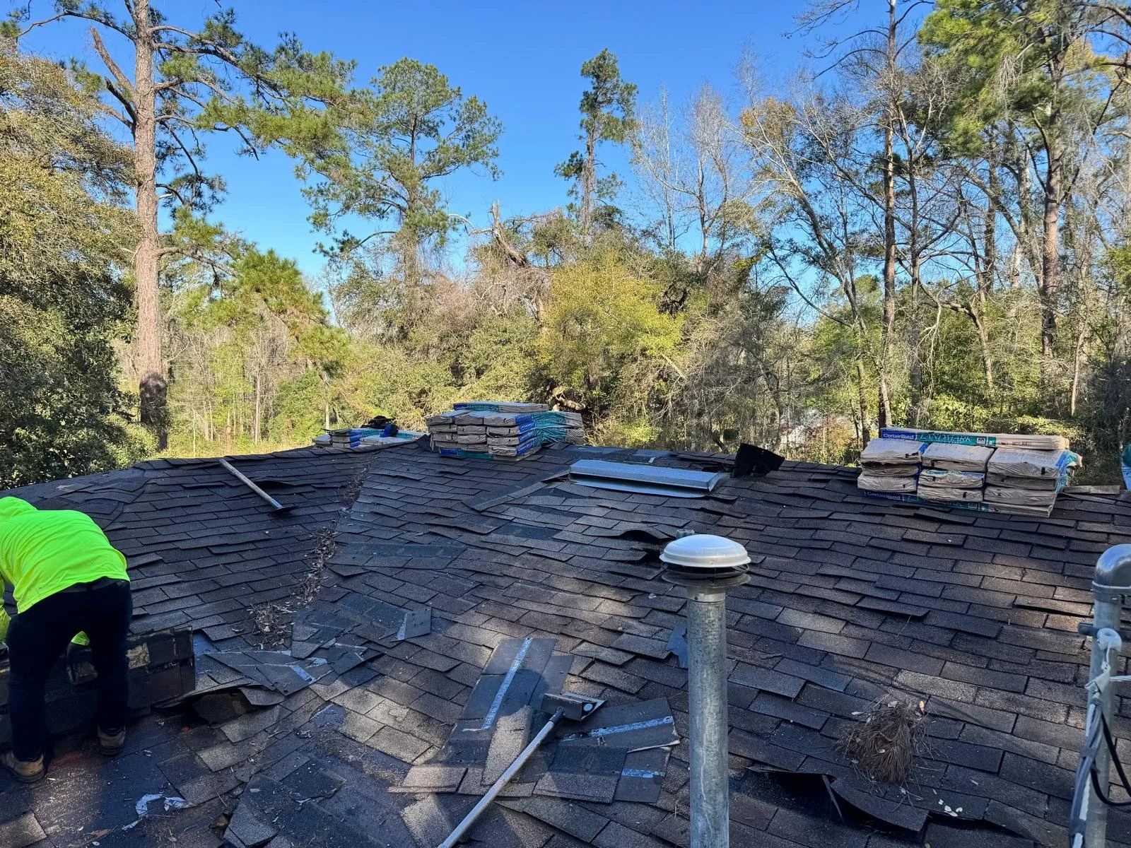 Roof construction scene with shingles, construction materials, and a worker in a neon yellow shirt, surrounded by a wooded area and blue sky.