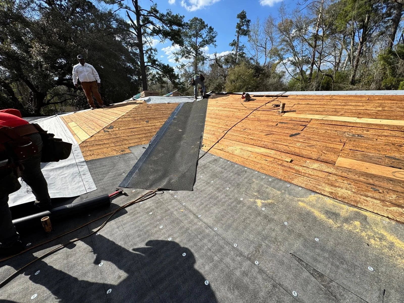 Three workers installing roofing shingles on a house under a clear blue sky, with trees visible in the background.