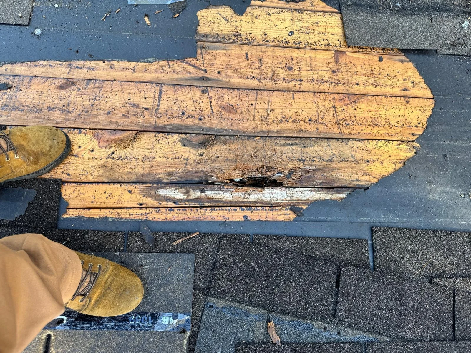 Damaged wooden planks on a roof with visible rot and decay, surrounded by black roofing tiles. Two yellow work boots are partially visible at the bottom of the image.