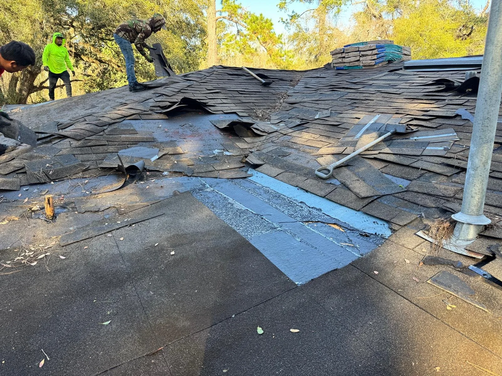 Roof being repaired with damaged shingles removed and new shingles laying on the roof, two workers present, one standing and one actively working, in a wooded area with trees in the background.
