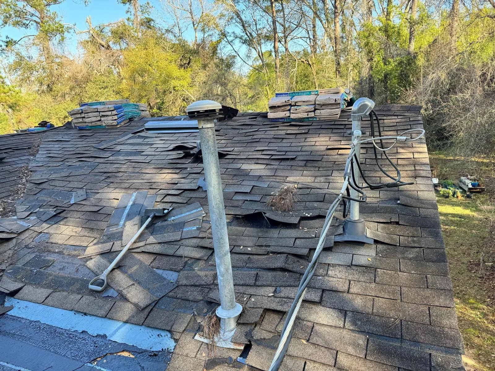 Photo of a roof with damaged shingles, roofing materials, a vent pipe, and electrical wires, with trees in the background.