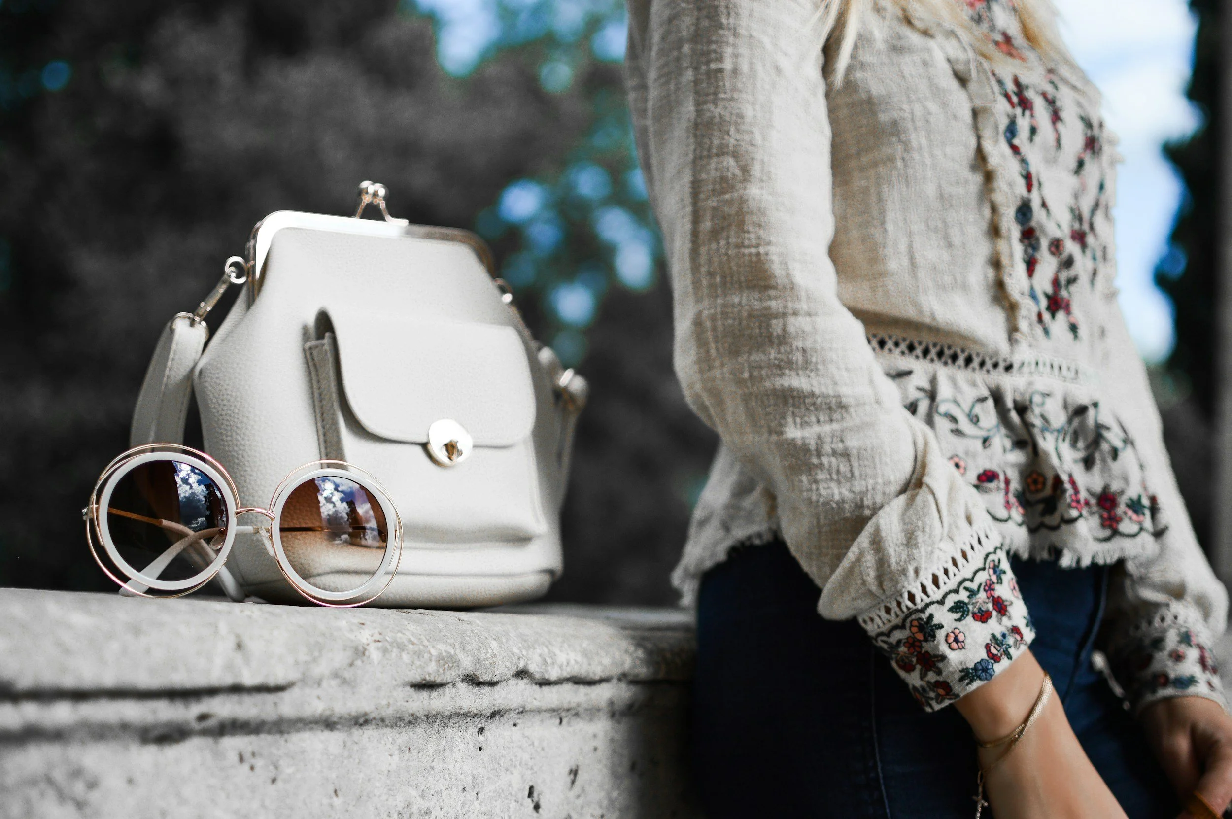 A white purse and a pair of round sunglasses with mirrored reflections are placed on a stone surface. A person wearing a cream-colored embroidered blouse with floral patterns is standing nearby, but their face is not visible.