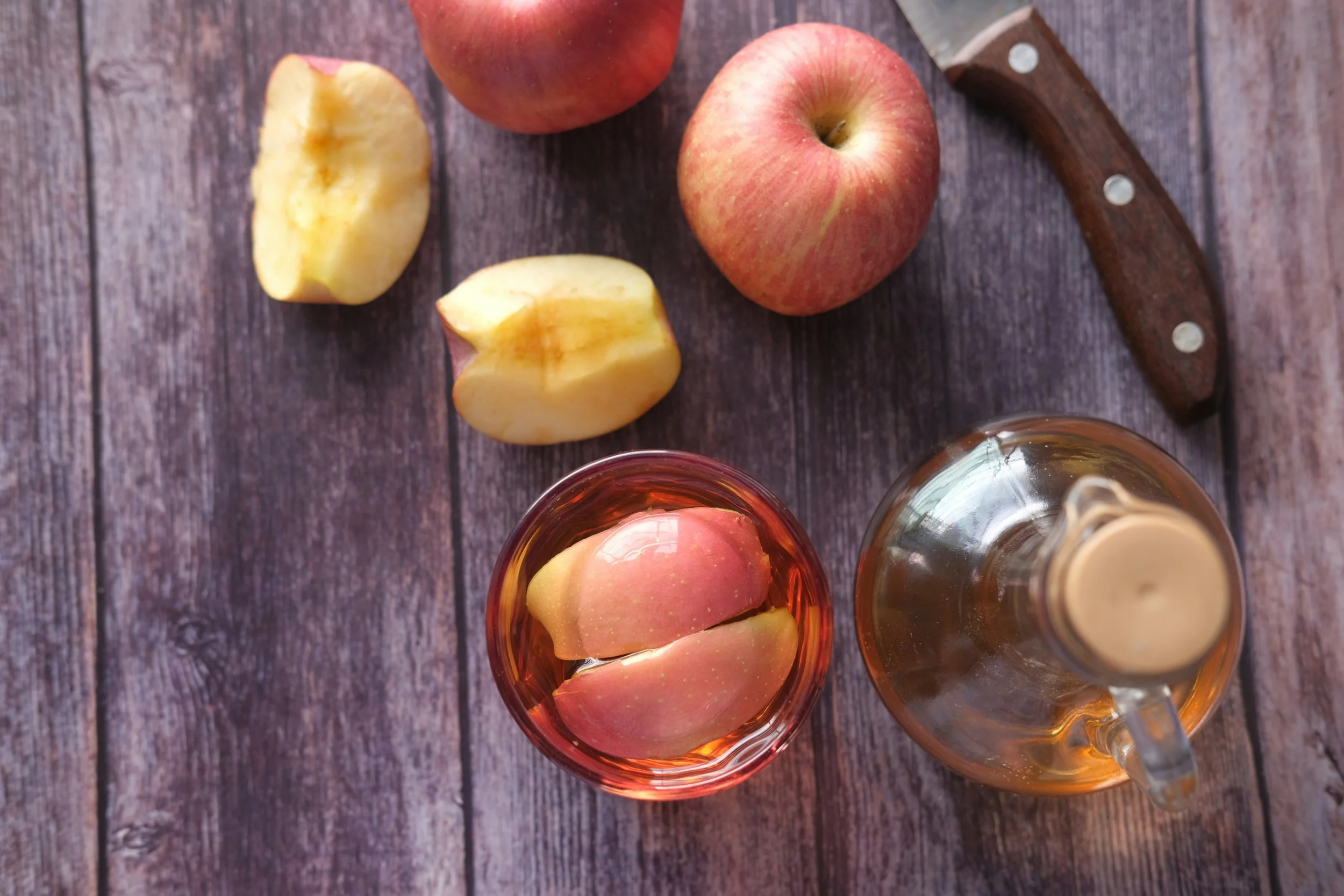 A wooden table with two apples and two apple slices, along with three glasses of drink, with one being a glass of apple juice.