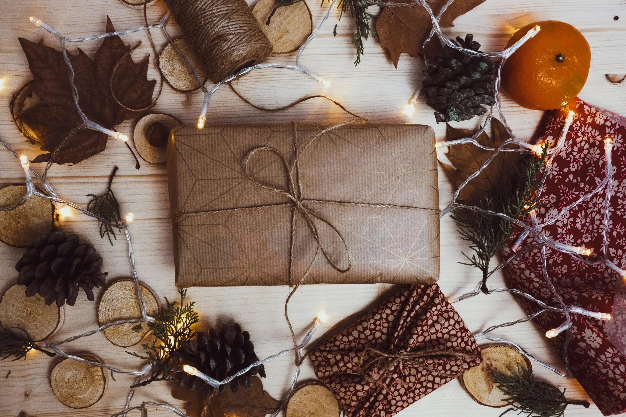 A flat lay of holiday decorations including wrapped gift, dried leaves, pinecones, sliced wood rounds, a small orange, and fairy lights on a light wooden surface.
