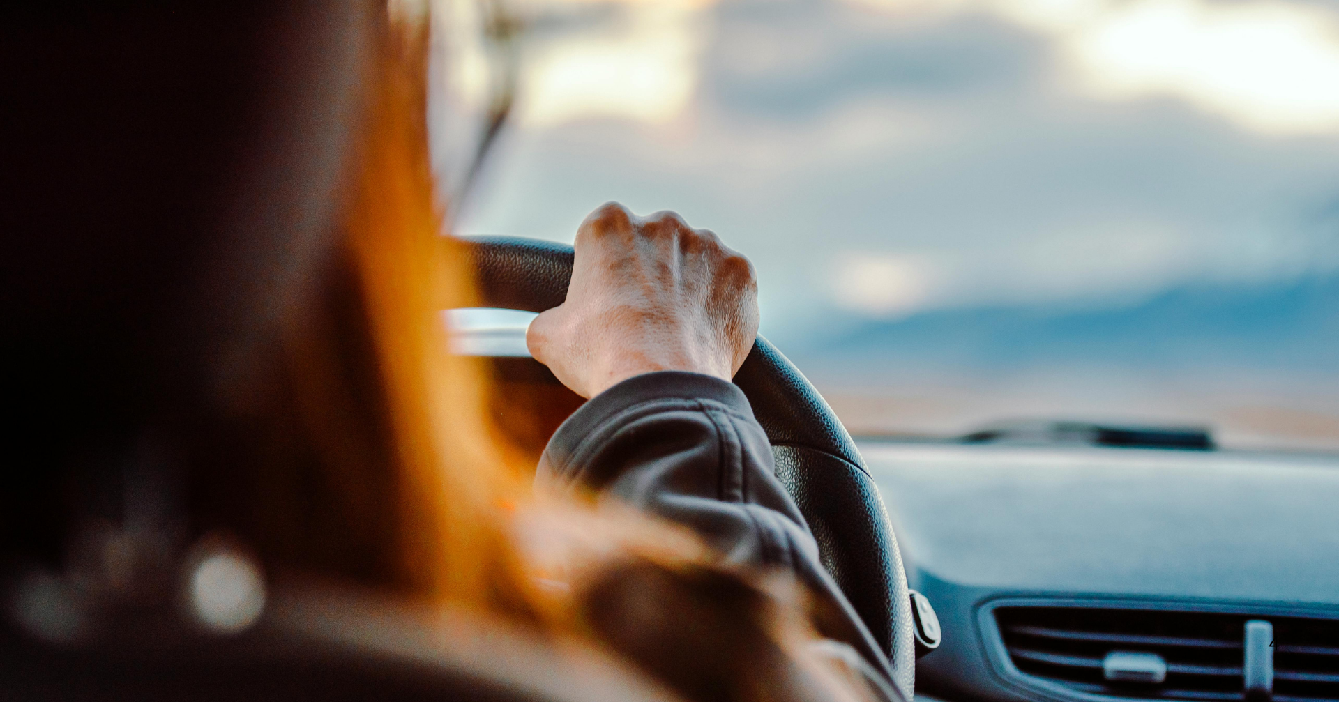 Person driving a vehicle, view of hand on steering wheel, blurred dashboard, and windshield showing a cloudy sky.