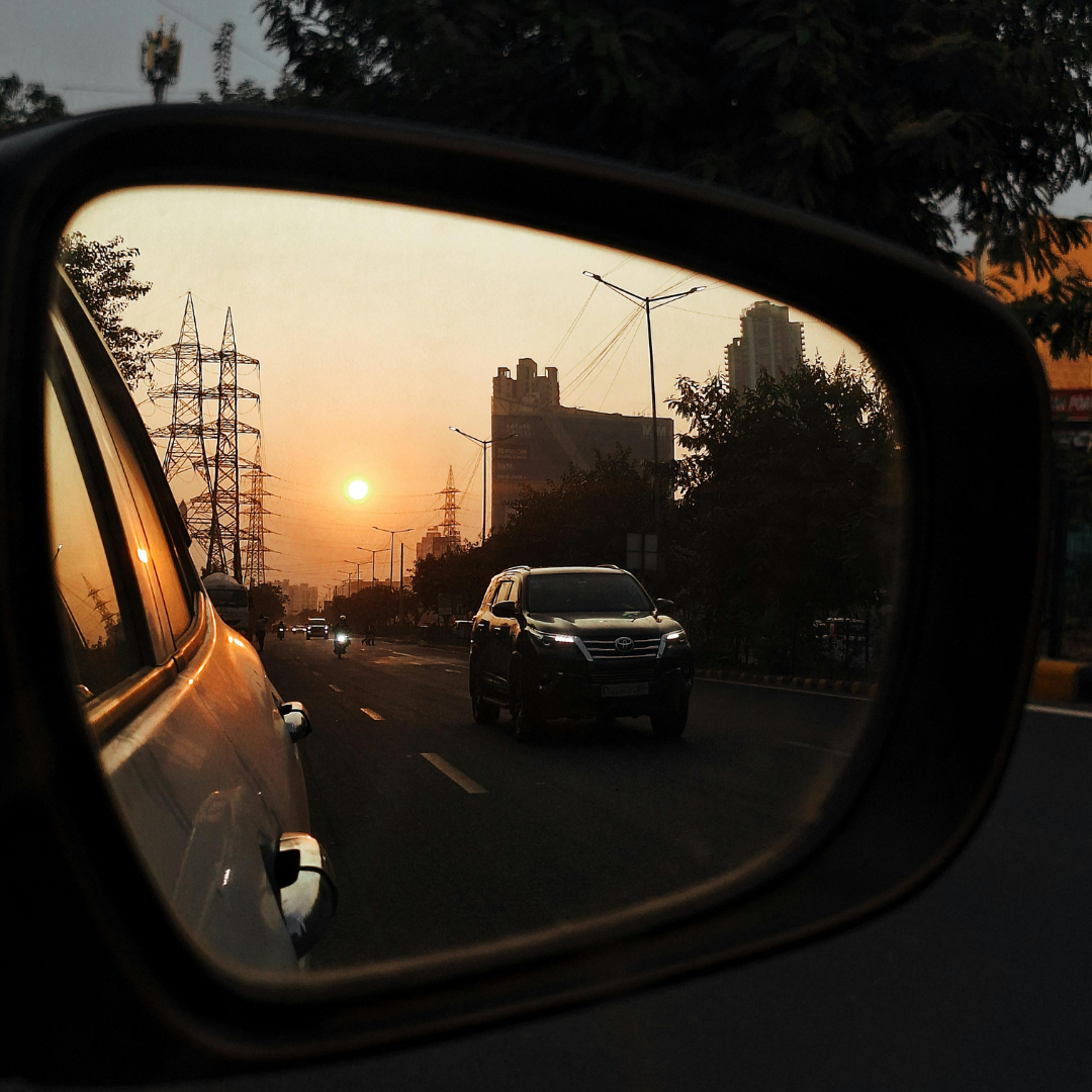 Sunset view captured through a car's side mirror, showing cars on the road, power lines, trees, and high-rise buildings in the distance.