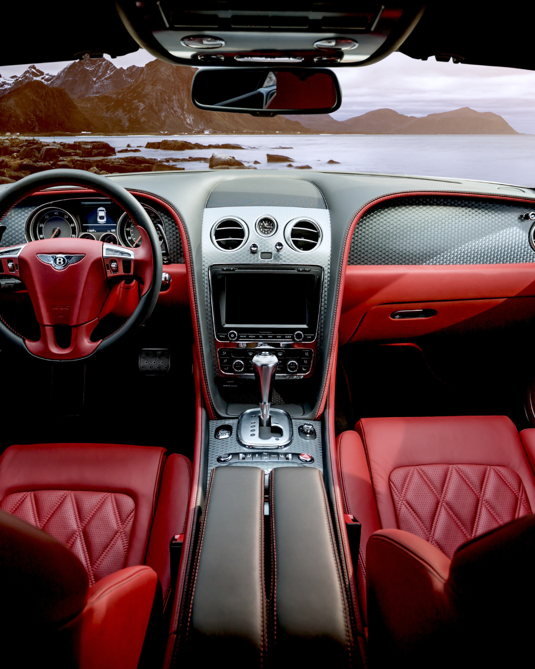Interior of a luxurious car with red leather seats, black accents, and a dashboard overlooking a scenic coastal landscape with mountains and rocks.