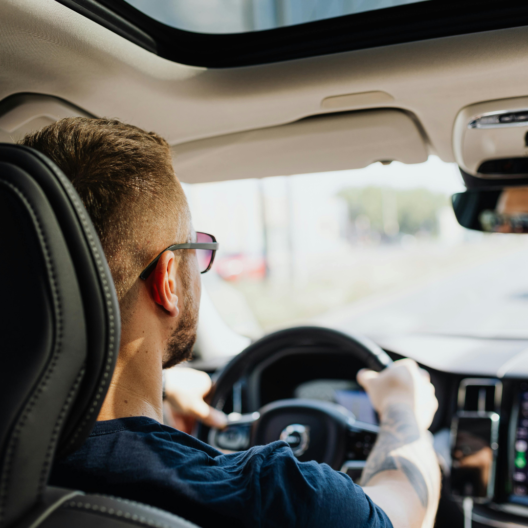 Man driving a vehicle, wearing sunglasses and a dark blue shirt, seen from behind inside the car.