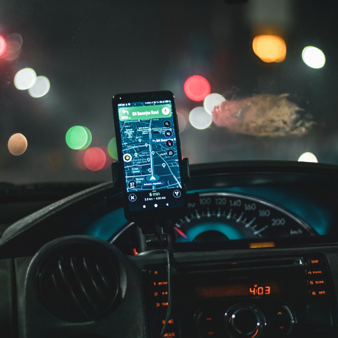 The image shows a GPS navigation app on a smartphone attached to a car dashboard, with the car's speedometer and dashboard clock displaying 4:03. Traffic lights are blurred in the background, indicating the photo was taken at night or in low light.