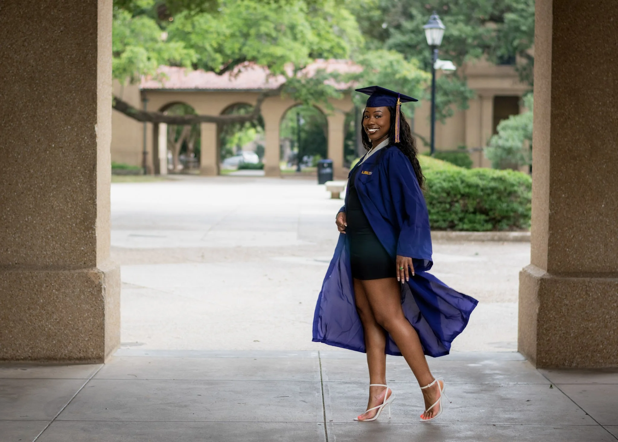 A young woman in a purple graduation gown and cap with tassel, smiling, standing under a stone archway, revealing her legs and white high-heeled shoes, with trees and a lamppost in the background.