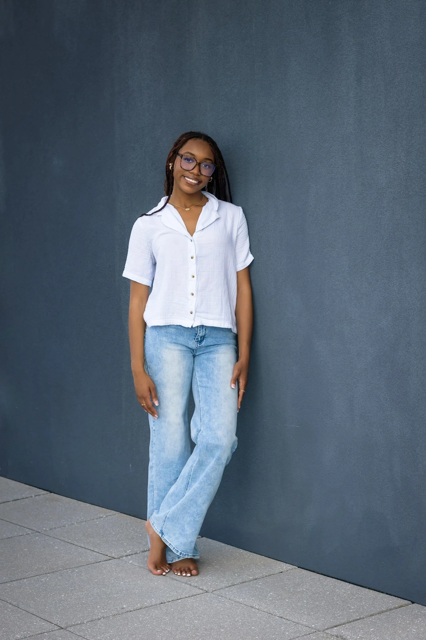 A woman with dark skin, wearing glasses, a white shirt, and light blue jeans, stands barefoot against a dark blue wall, smiling and leaning slightly on the wall.