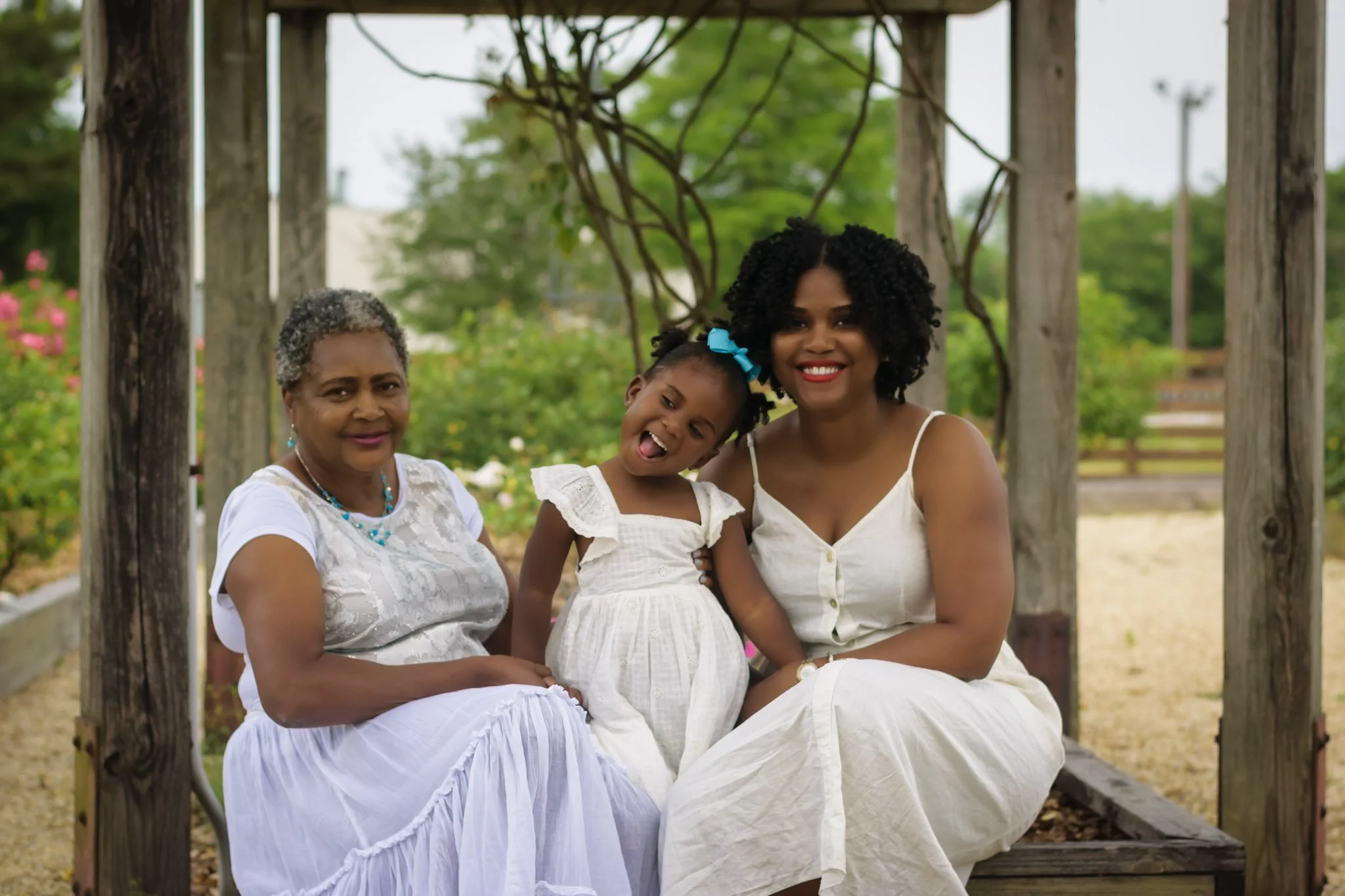 Three women and a young girl sitting together on a wooden outdoor bench with greenery in the background, smiling and enjoying each other's company.