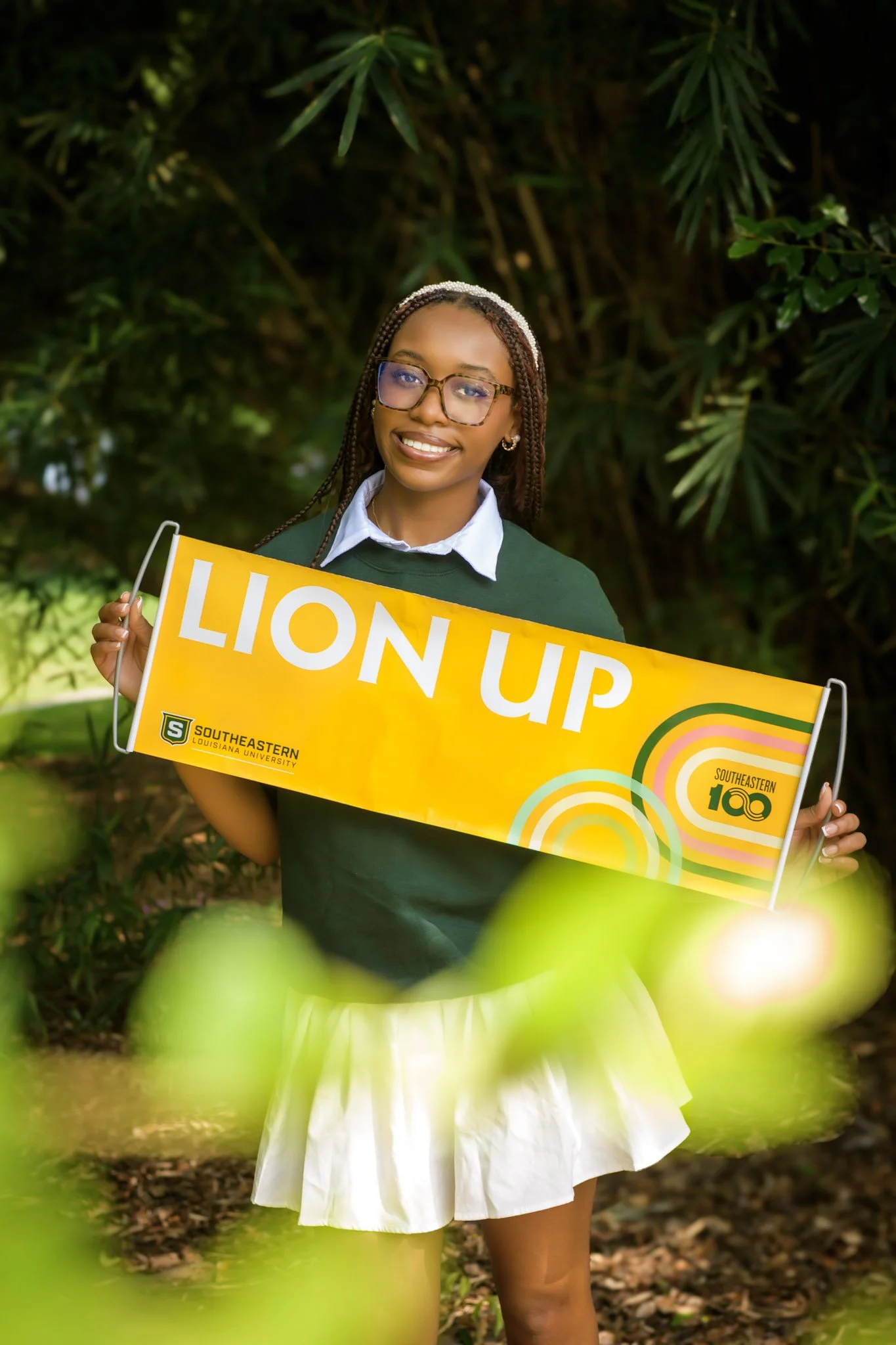 A young woman with braided hair, glasses, and earrings holds a yellow celebration sign that reads 'Lion Up' with Southeastern Louisiana University logo, standing outdoors among green foliage.