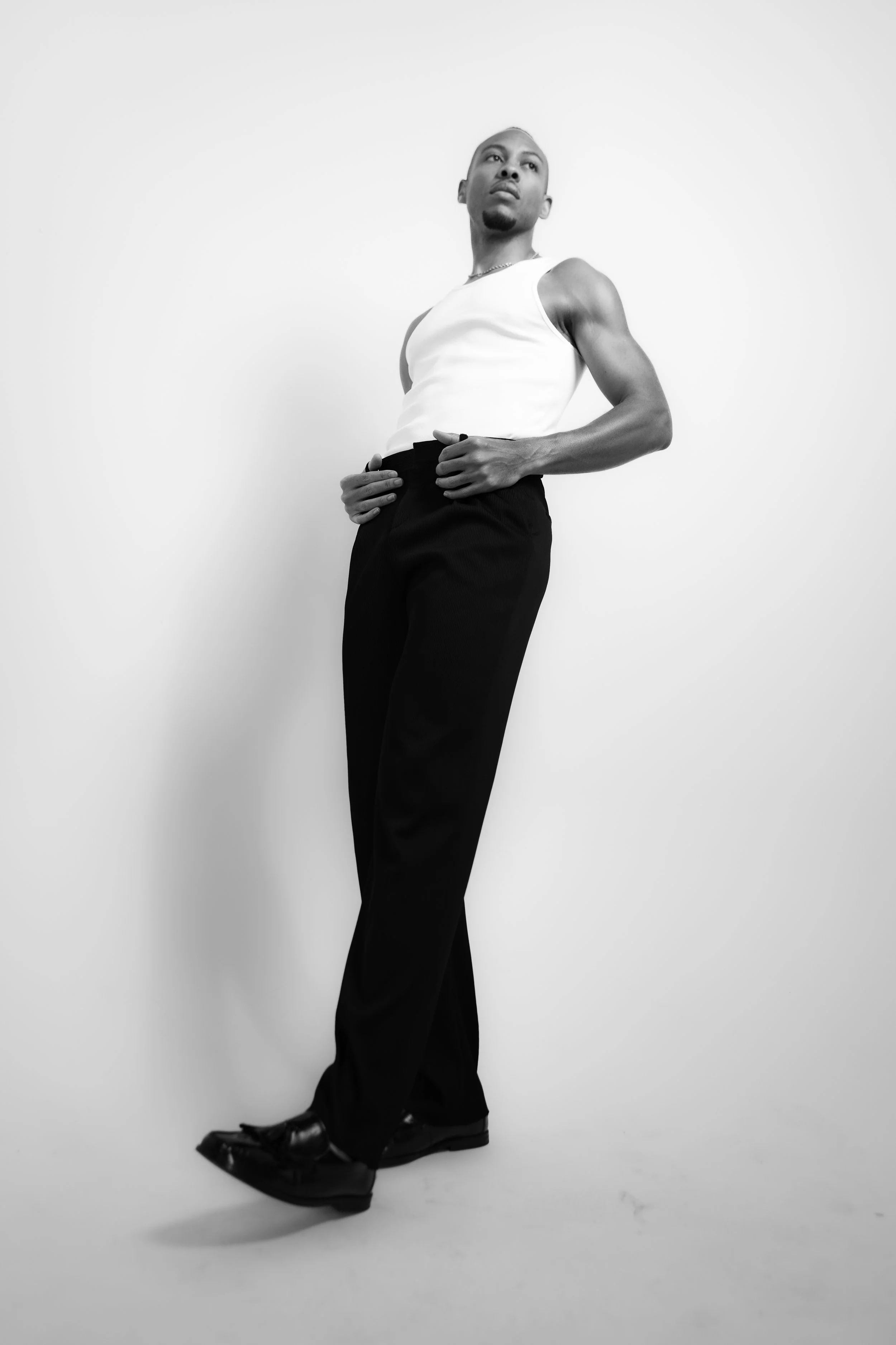 A black and white photo of a man standing against a plain light-colored wall, wearing a sleeveless white shirt, black pants, and black shoes, looking off to the side with a confident pose.