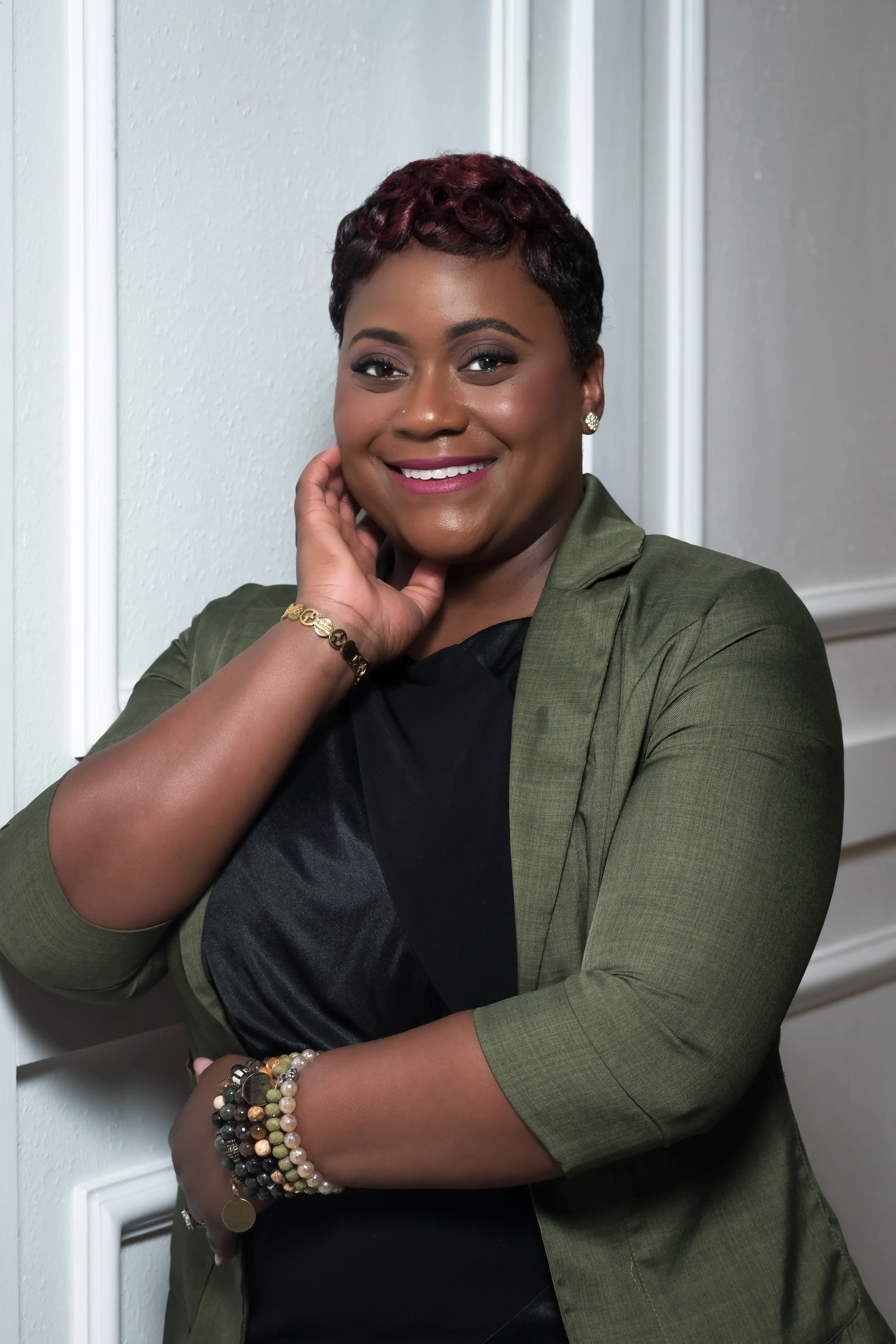 Portrait of a smiling African American woman with short curly hair, wearing earrings, a green blazer, and beaded bracelets on her wrist, posed with her hand near her face against a neutral background.