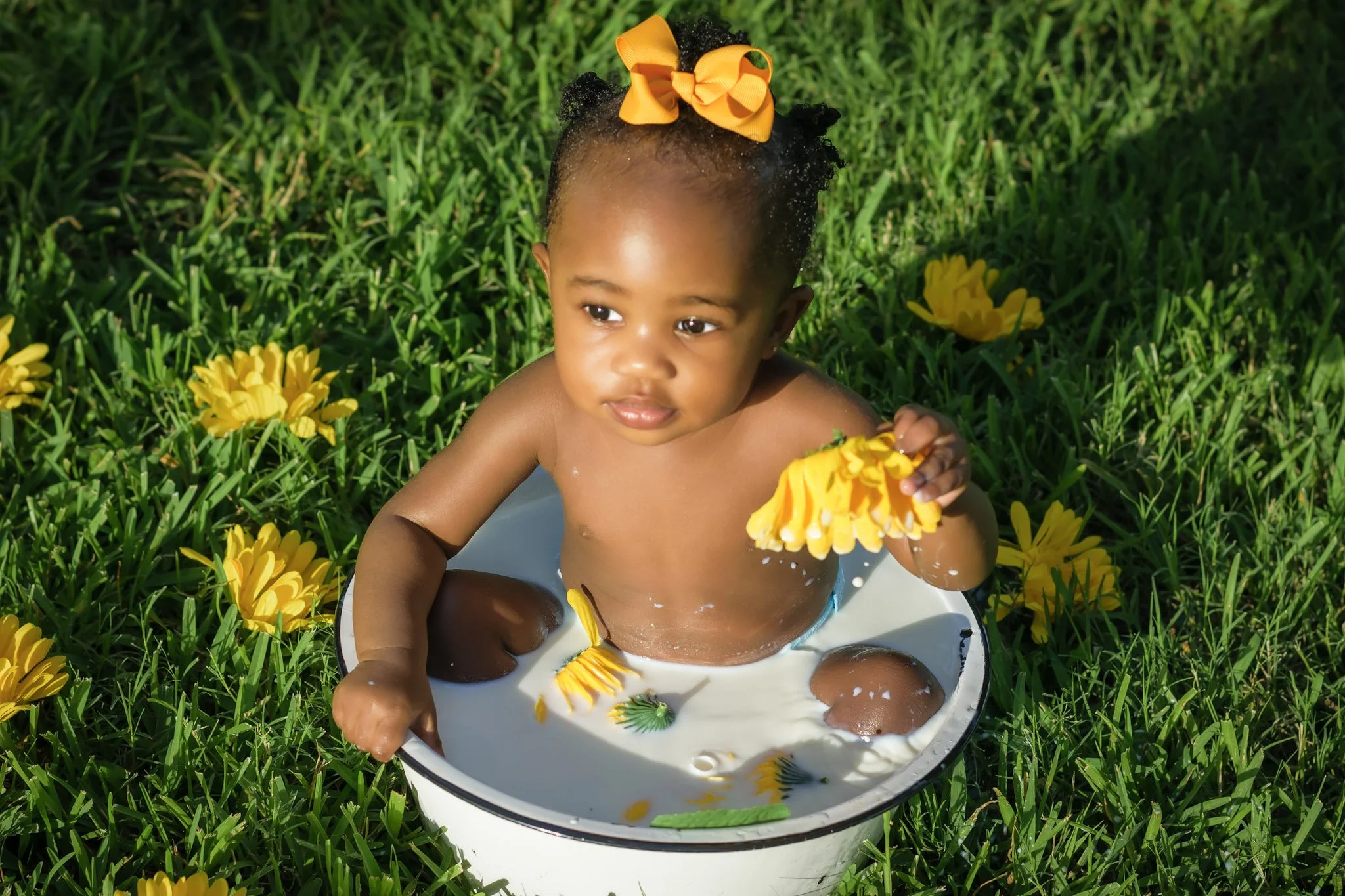 A young girl with curly hair and a yellow bow sits in a shallow bowl filled with water and yellow flowers, surrounded by green grass and yellow flowers outdoors.