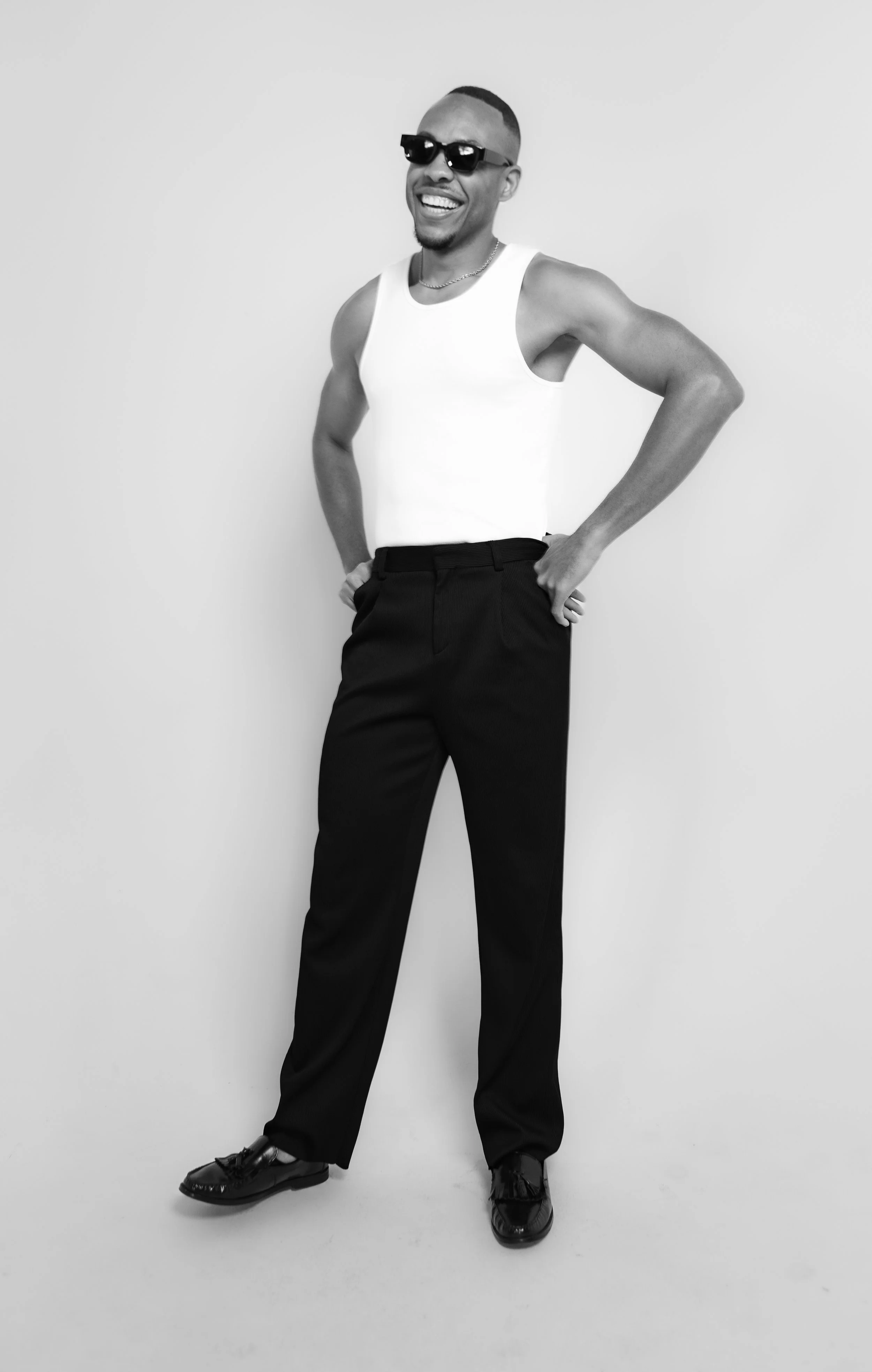 A man smiling and wearing sunglasses, a white tank top, black pants, and dress shoes, standing against a plain light-colored background.