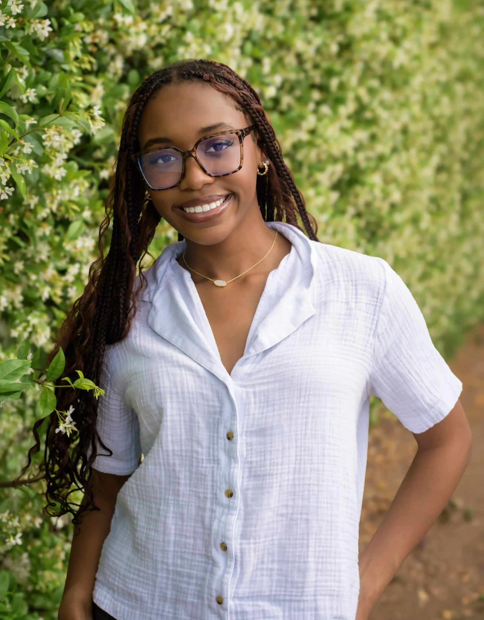 A young woman with braids and glasses smiling outdoors, standing next to green bushes with small white flowers.