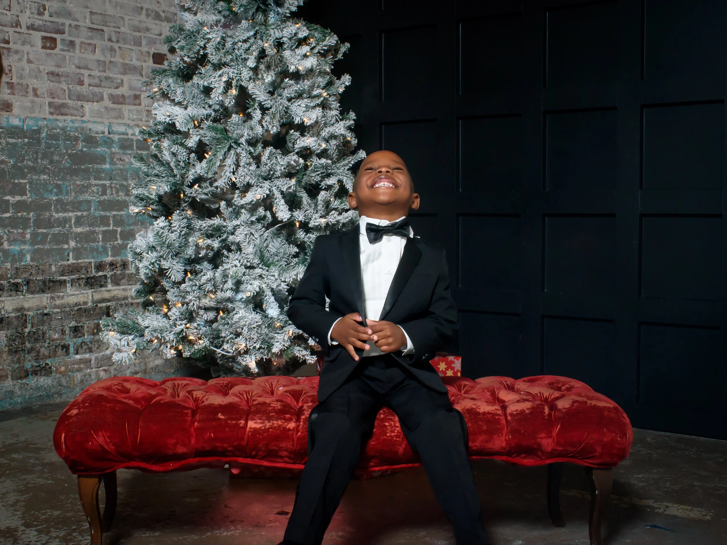 A young boy in a tuxedo smiling while sitting on a red velvet velvet tufted bench in front of a decorated Christmas tree, with a brick wall and dark wall behind him.