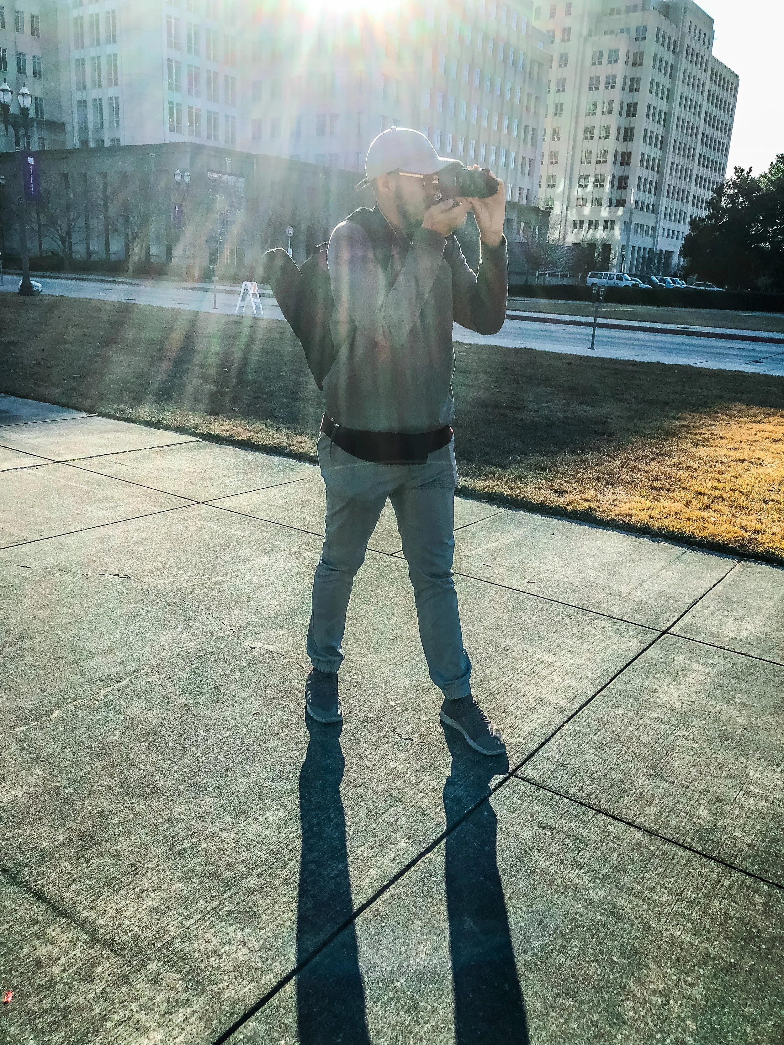 Person taking a photograph with a camera in an outdoor urban area, standing on a sidewalk with buildings in the background, sunlight creating a lens flare.
