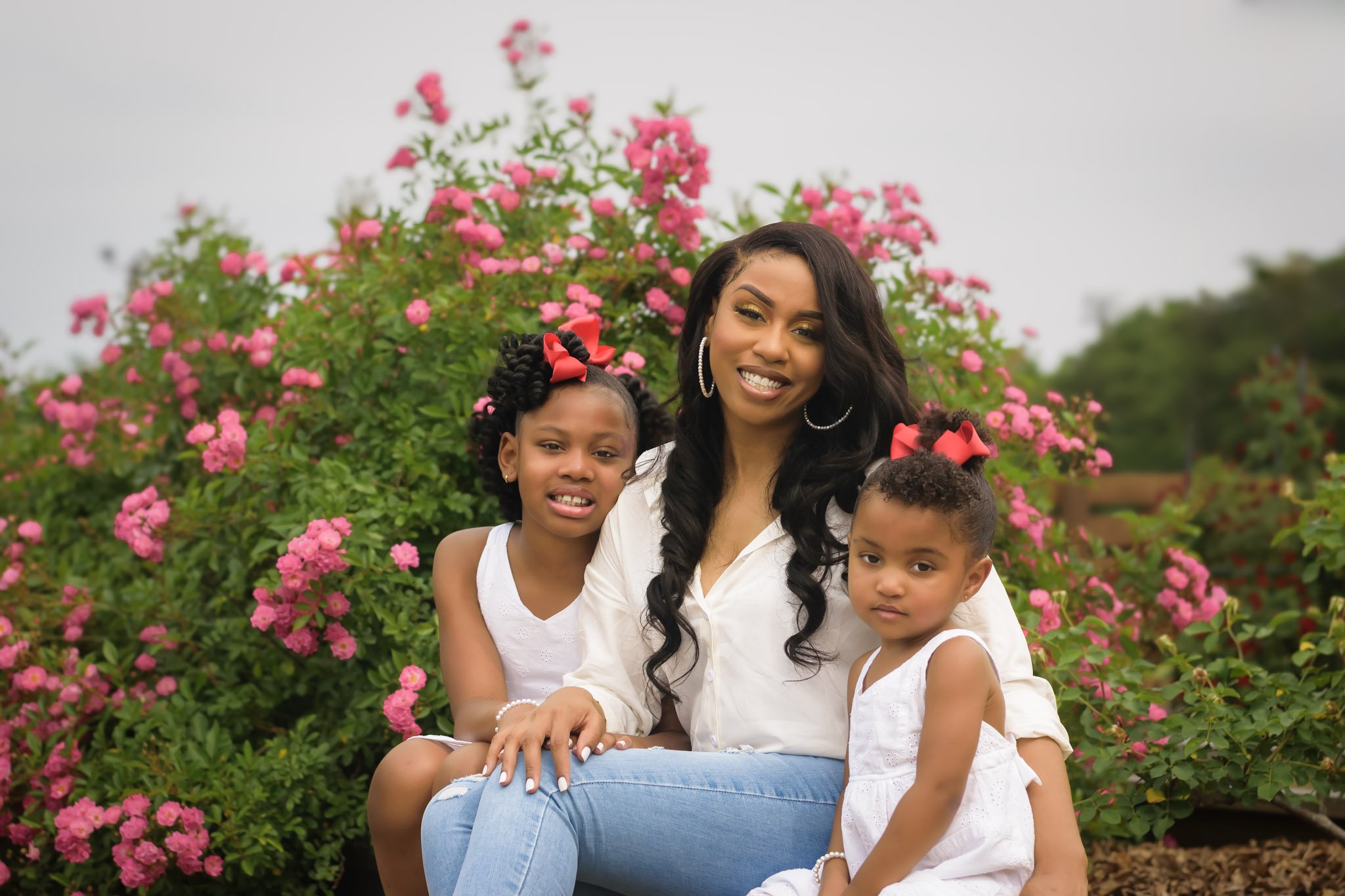 Smiling woman with long dark hair and hoop earrings sitting outdoors with three young girls near pink flowering bushes.