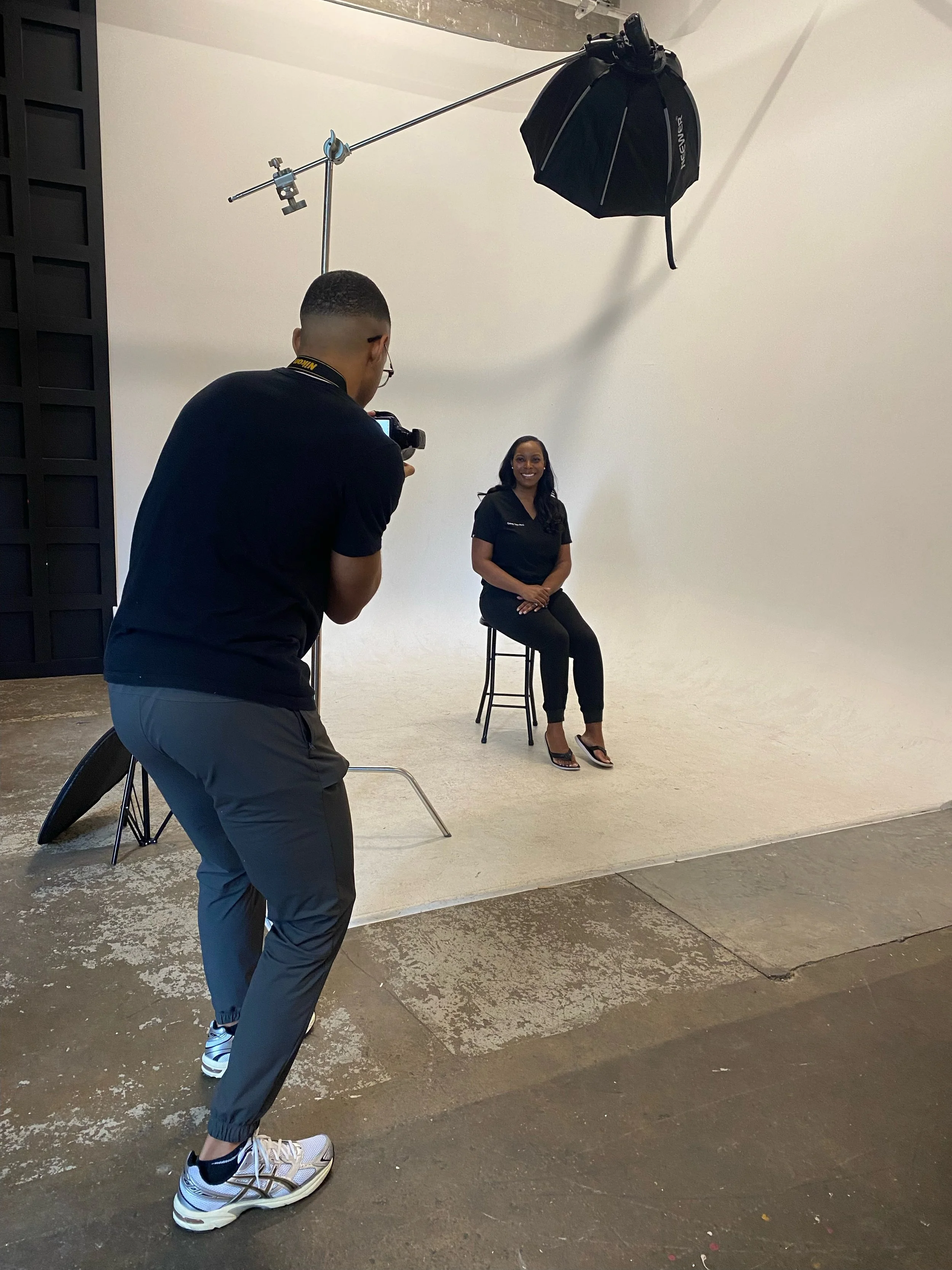 A woman sitting on a stool in a photography studio, smiling at the camera. A man in casual clothing is taking her photo. Studio equipment includes a large softbox light, a black reflector, and a white backdrop.
