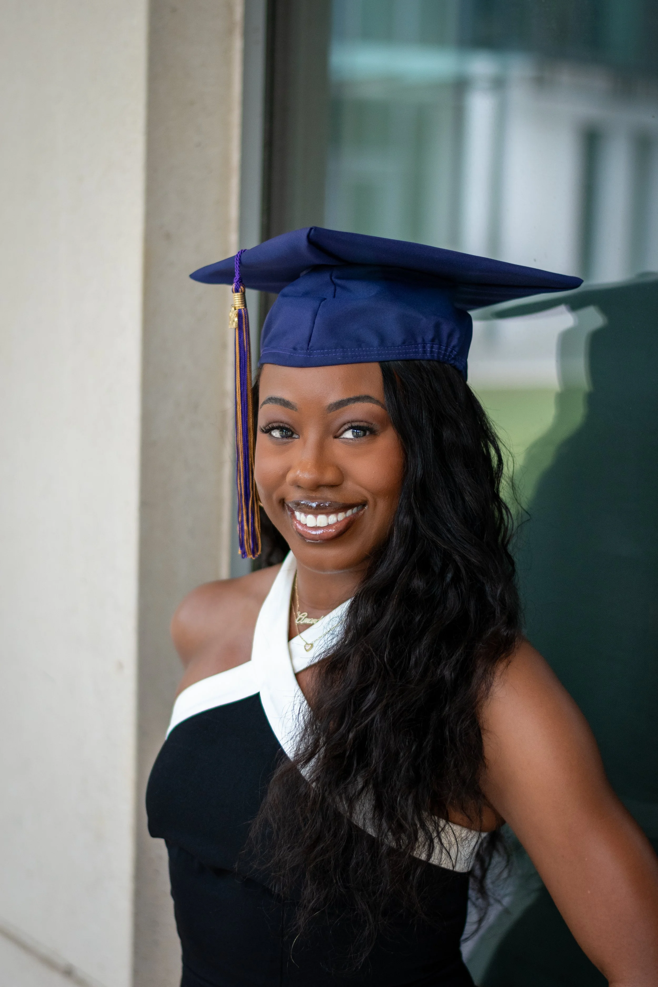 A young woman with long, dark, wavy hair wearing a blue graduation cap and a black dress with a white neckline smiling at the camera.