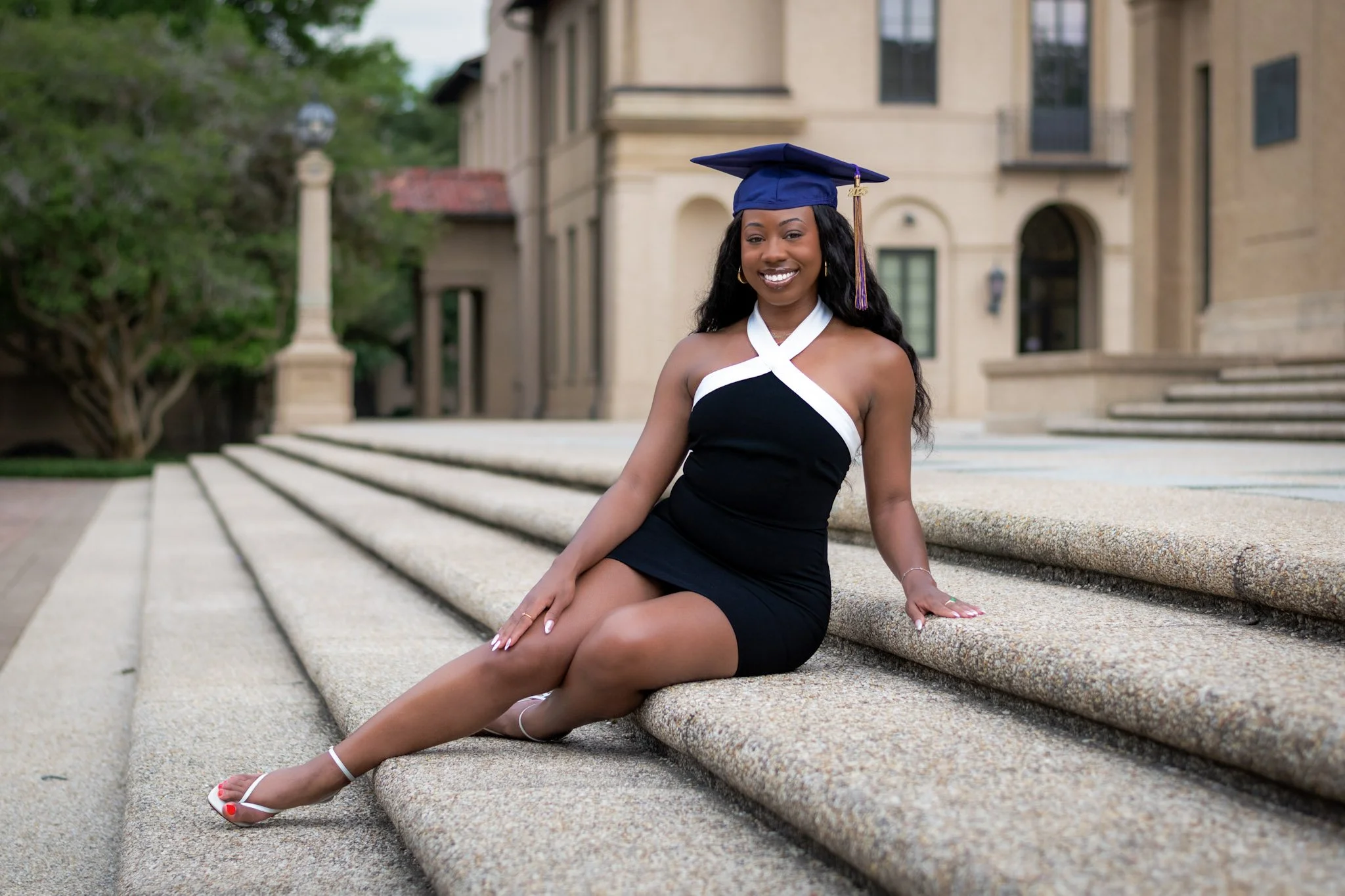 Young woman in black and white dress with a graduation cap sitting on stone stairs outside a building.