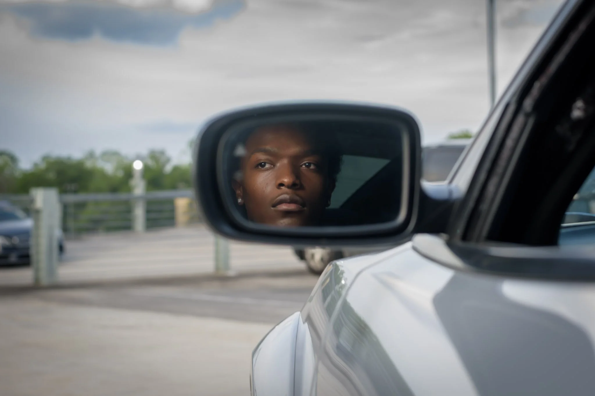 The image shows a reflection of a woman in the side mirror of a car, with a cloudy sky and some trees in the background.