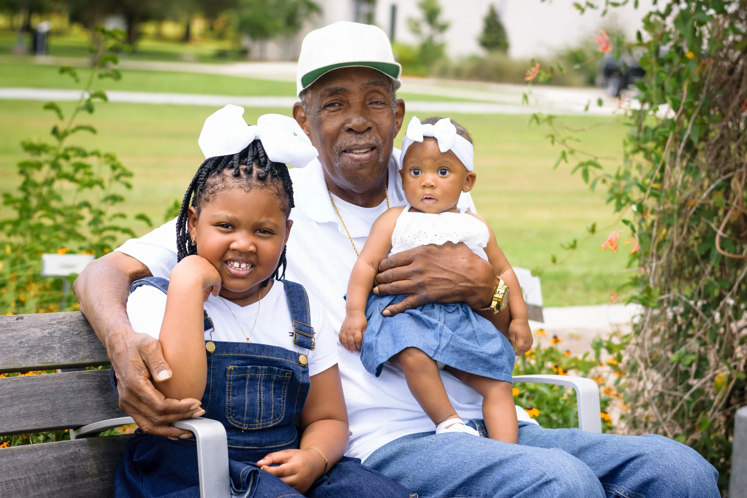 An elderly man sitting on a park bench holding two young children, one girl and one baby, with greenery and flowers in the background.