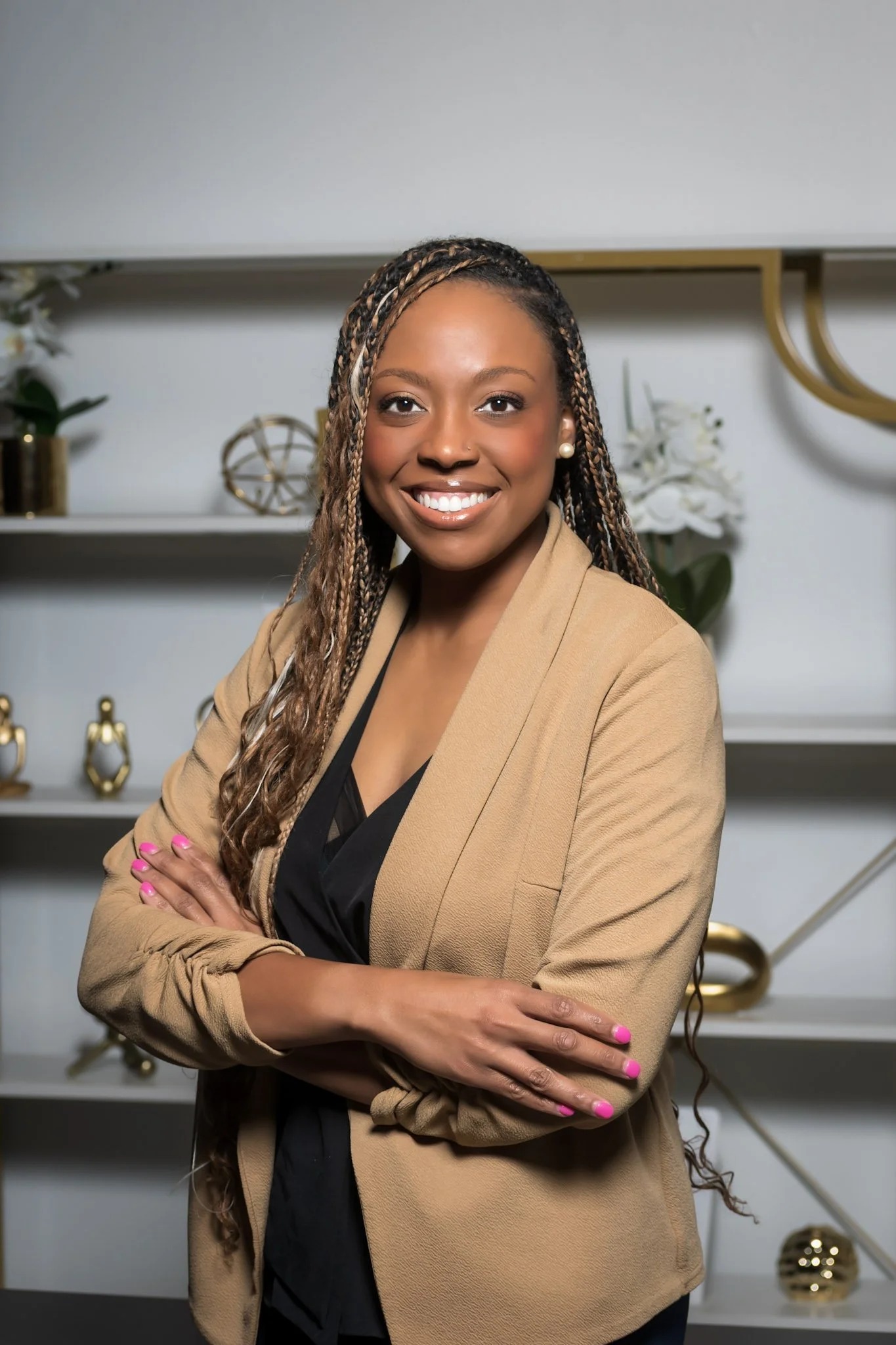 A smiling woman with long braided hair, wearing a tan blazer and black top, standing with arms crossed in front of a modern shelf decorated with gold and white ornaments and floral arrangements.