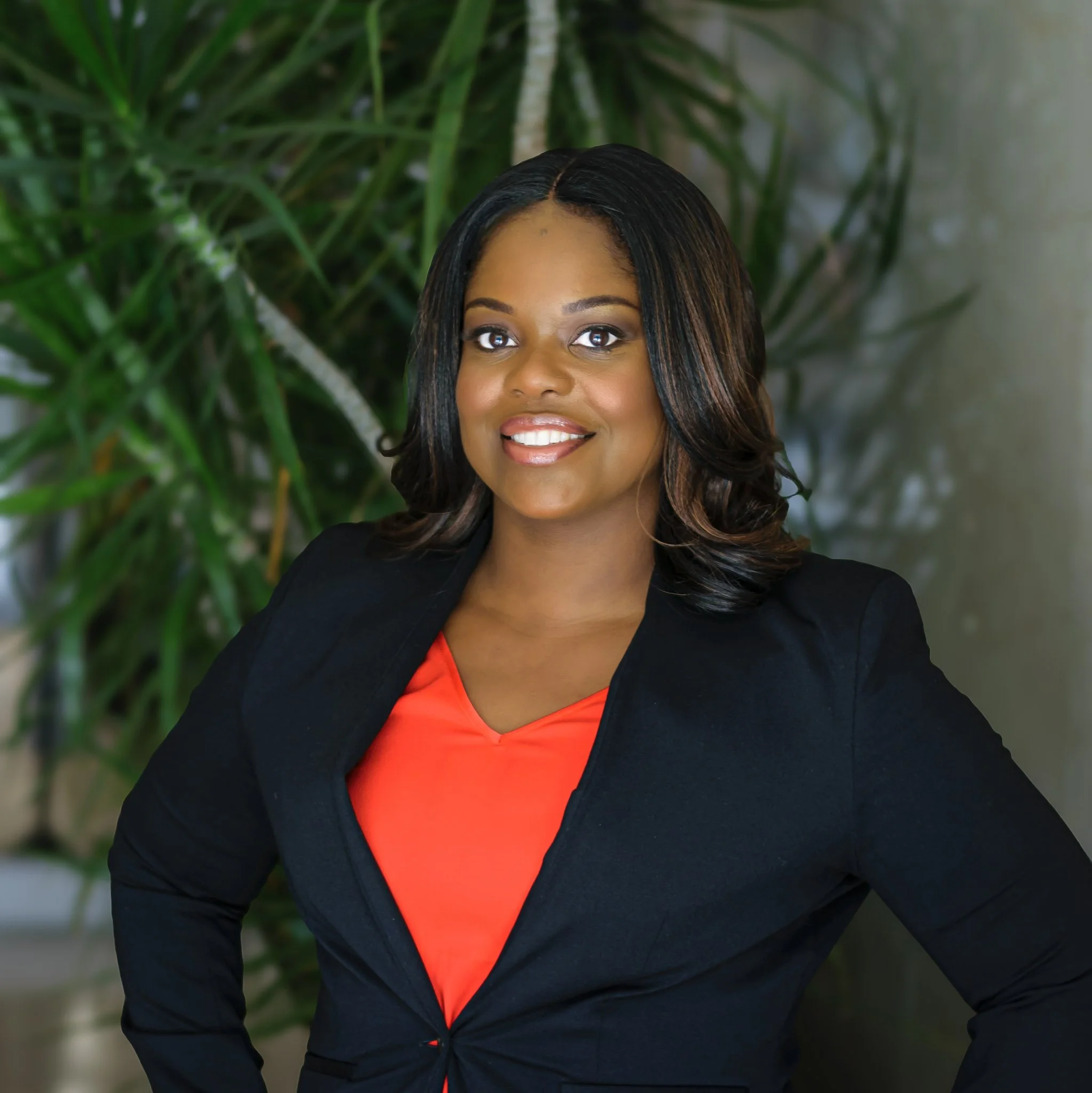 Young African American woman smiling, wearing black blazer over red top, standing outdoors with green foliage in the background.
