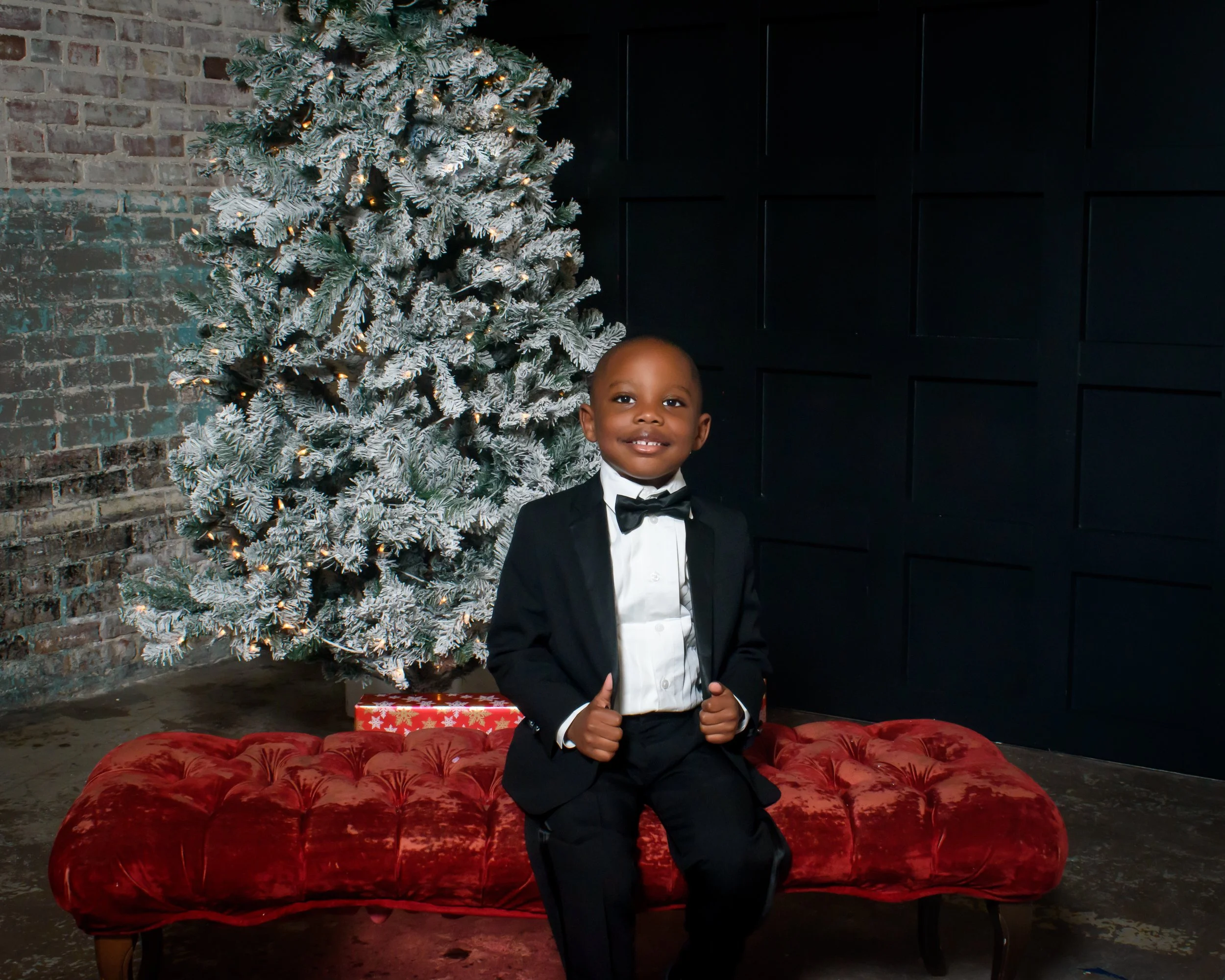 A young boy in a black tuxedo and bow tie, sitting on a red velvet tufted bench, giving a thumbs-up, with a decorated Christmas tree with white snow-like frosting and lights in the background, and a wrapped Christmas gift on the floor.