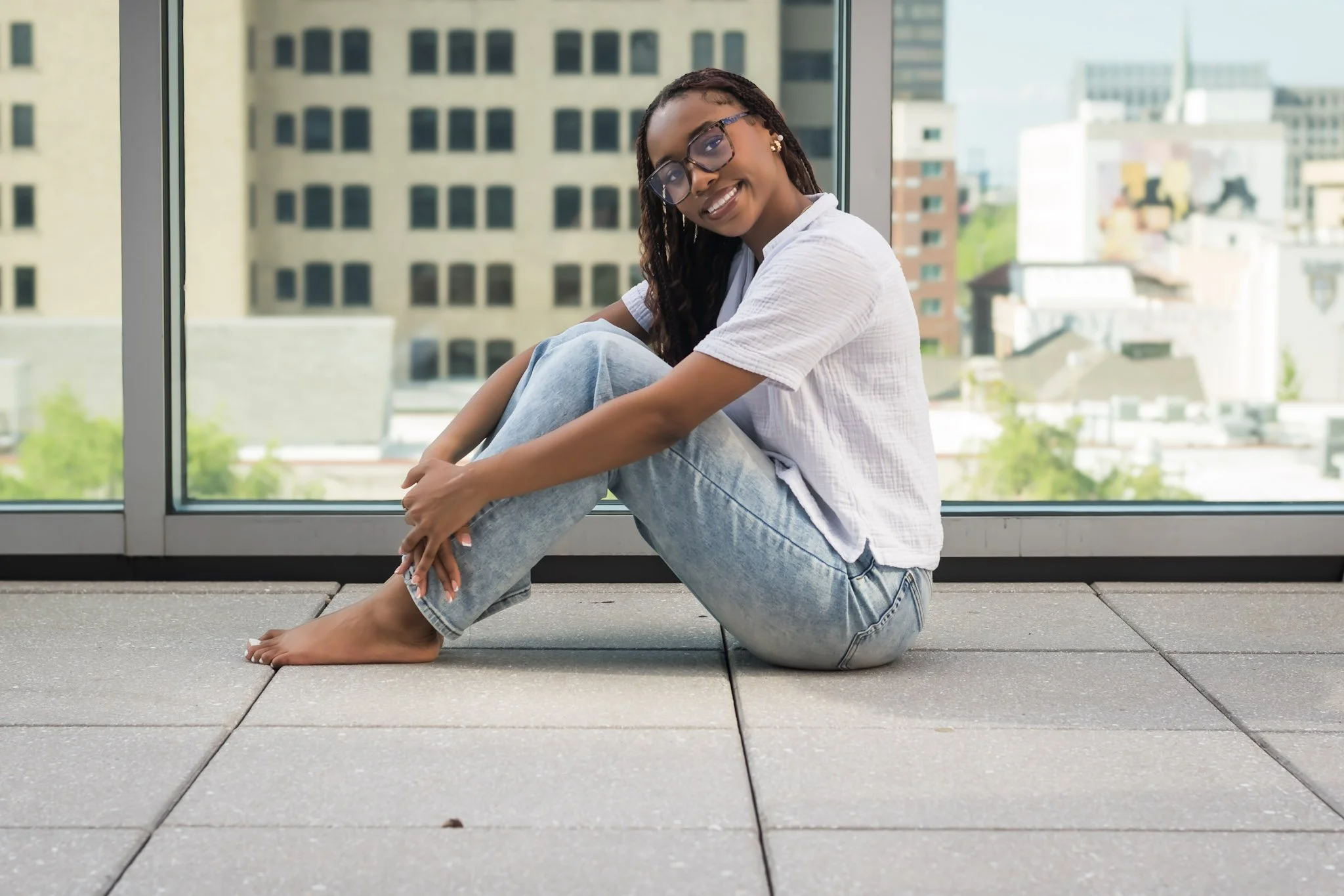 Young woman with glasses sitting barefoot on the floor near a large window in an urban setting, smiling at the camera.