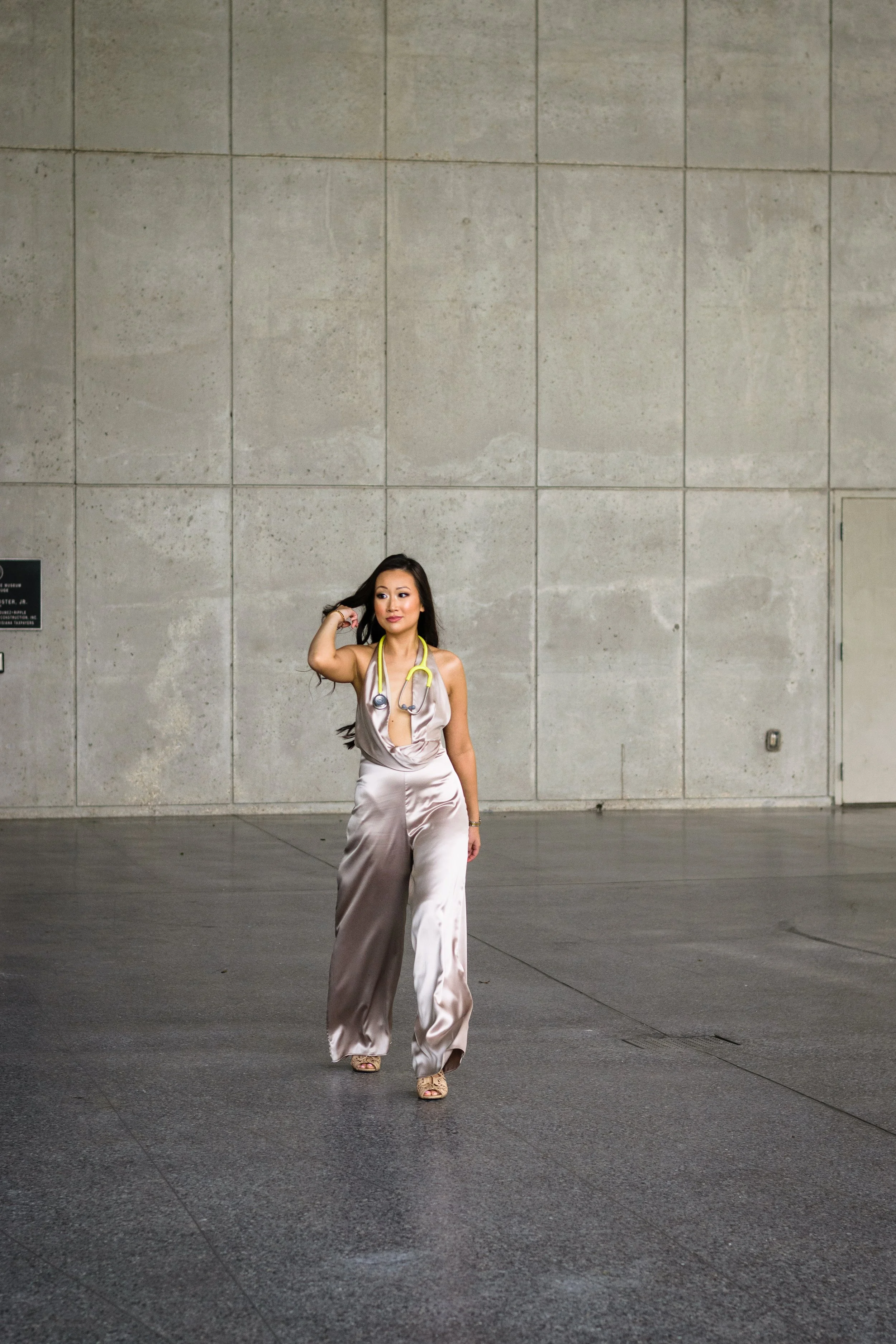 A woman in satin beige pants and top with a stethoscope around her neck walking on a paved sidewalk in front of a concrete wall.
