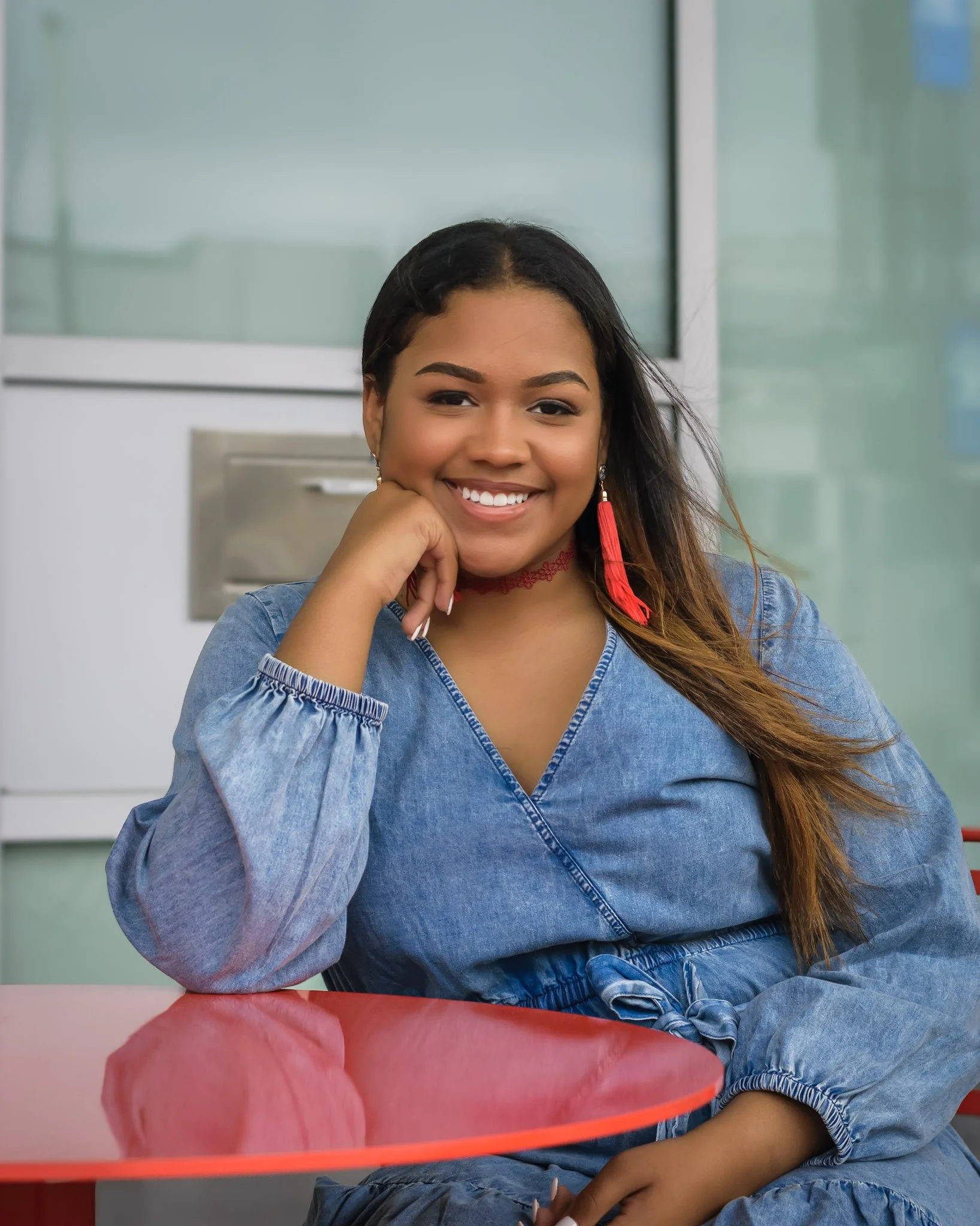 Young woman sitting at a red table, smiling, with hand resting on her chin, wearing a denim jacket and red earrings, in a modern indoor setting.