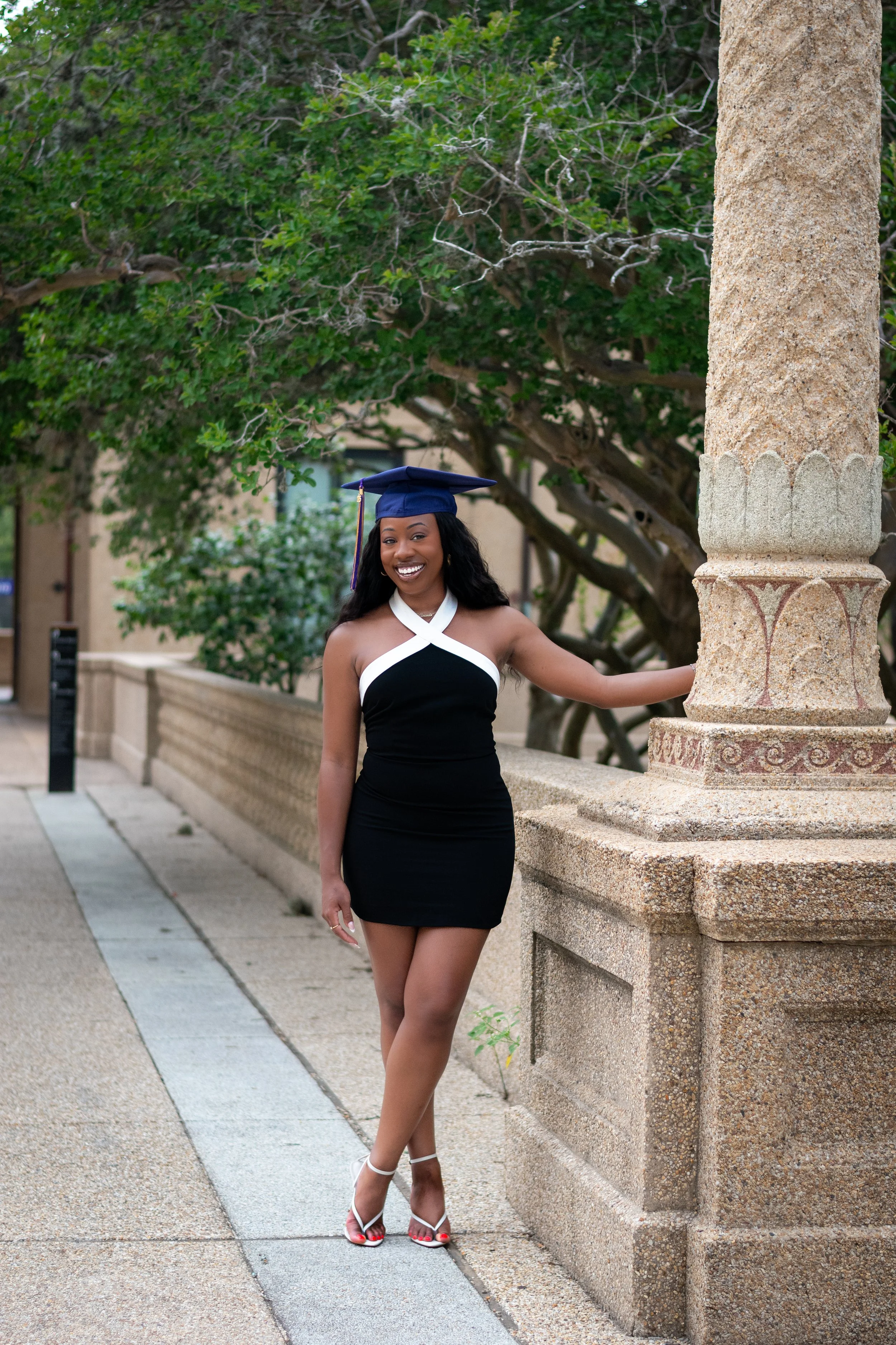Young woman wearing a black and white dress, white heels, and a blue graduation cap with a tassel, standing outdoors near a stone railing and leafy trees.