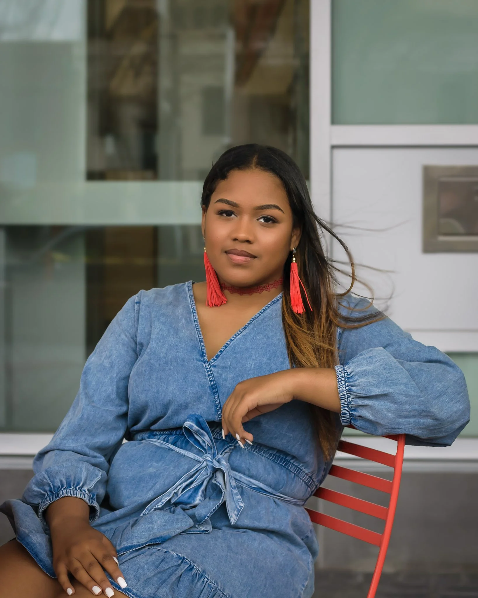 A young woman sitting on a red bench outdoors, wearing a denim outfit, red tassel earrings, and a red lace neck accessory, with a blurred modern glass building in the background.