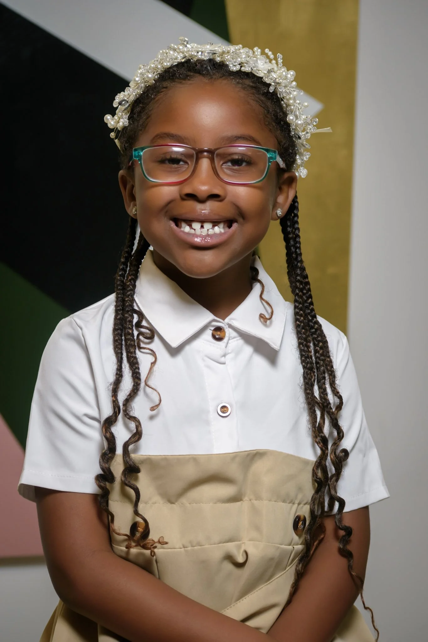 A young girl with long, braided hair, wearing glasses, a decorative pearl headband, and a white blouse with a beige apron, smiling.