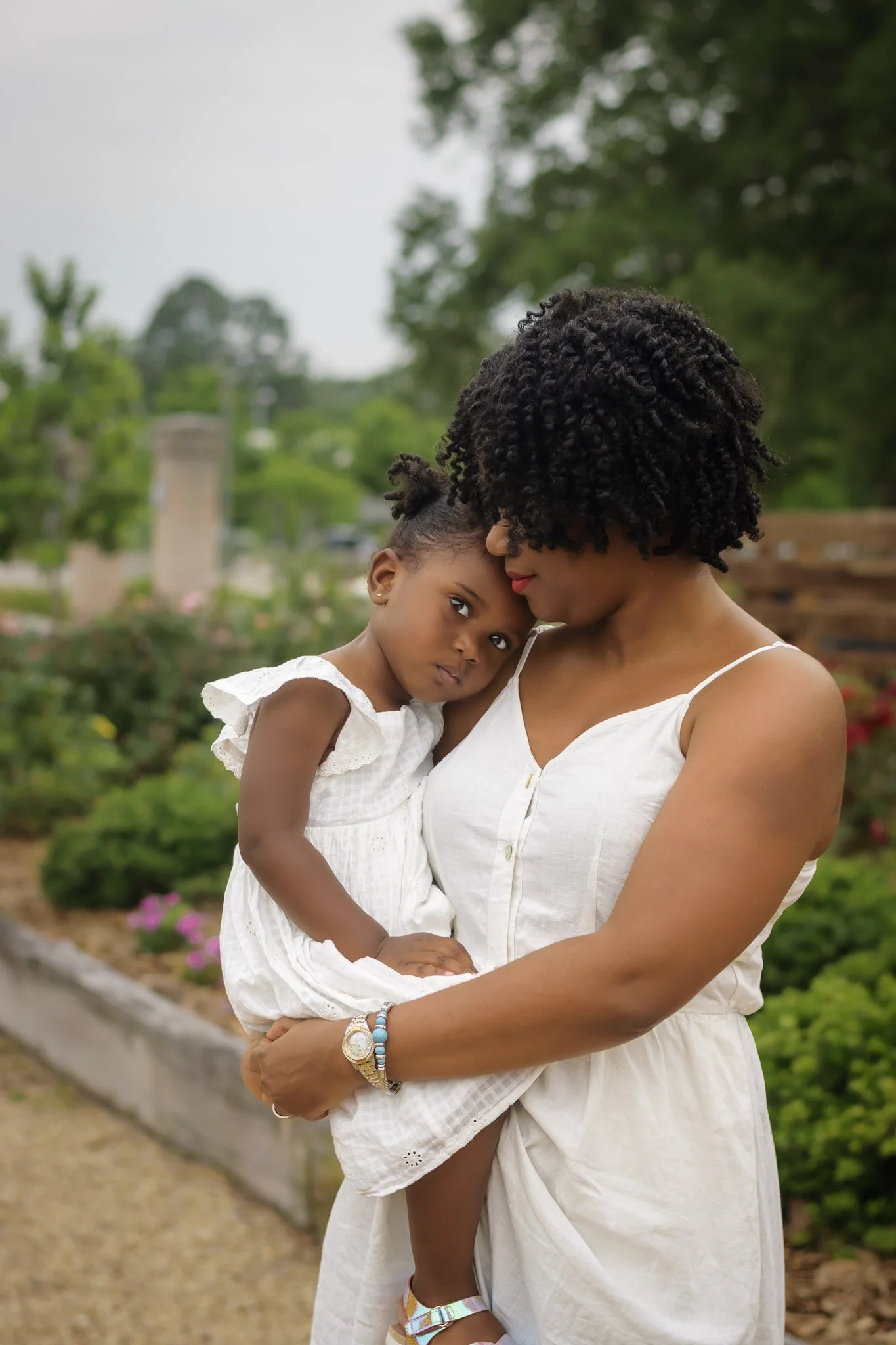 A woman holding a young girl in a garden with trees and flowers in the background. Both are wearing white dresses and have dark curly hair.