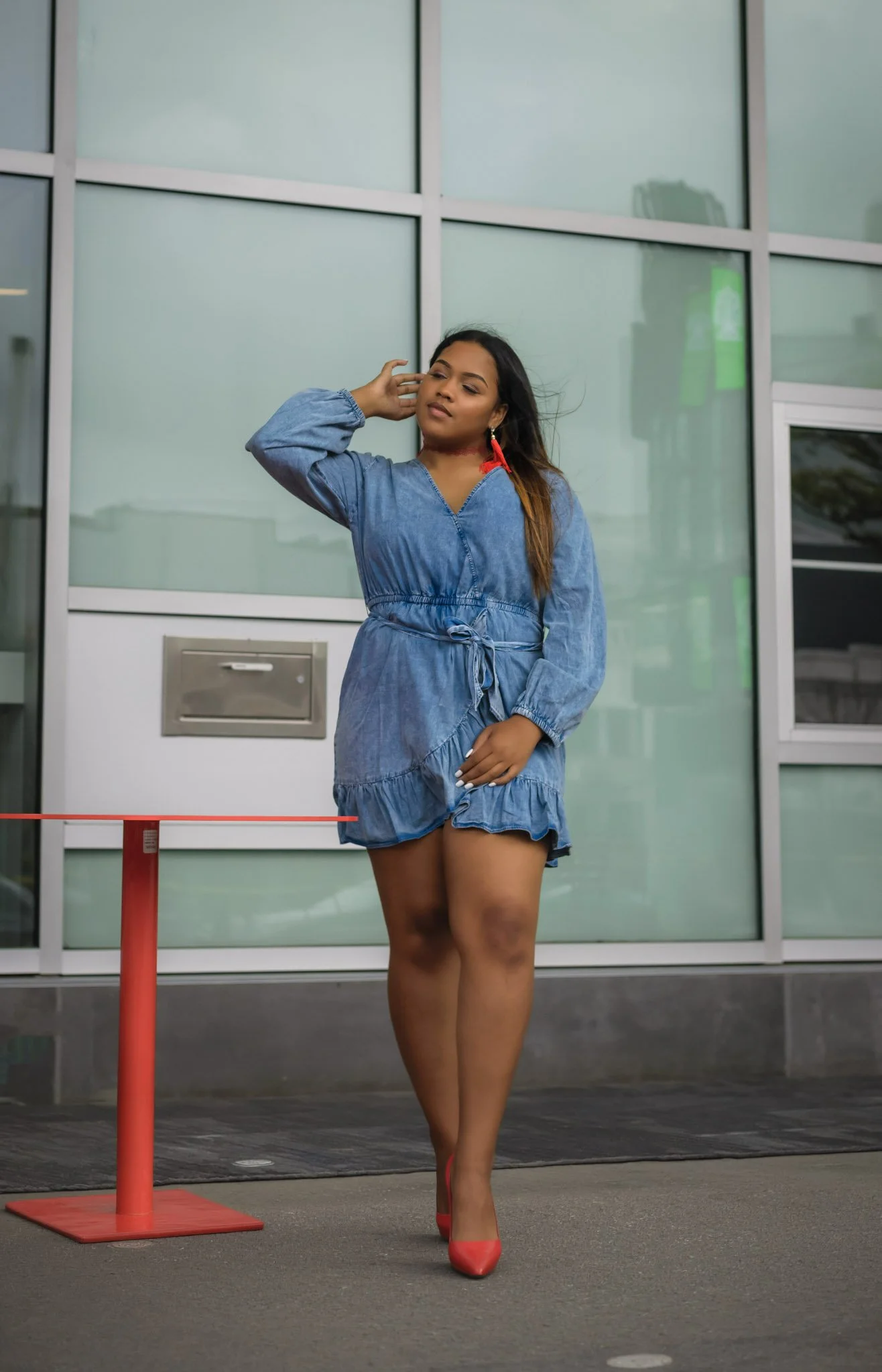 A woman standing outside near a glass building, wearing a blue denim dress and red high heels, with her right hand touching her head.
