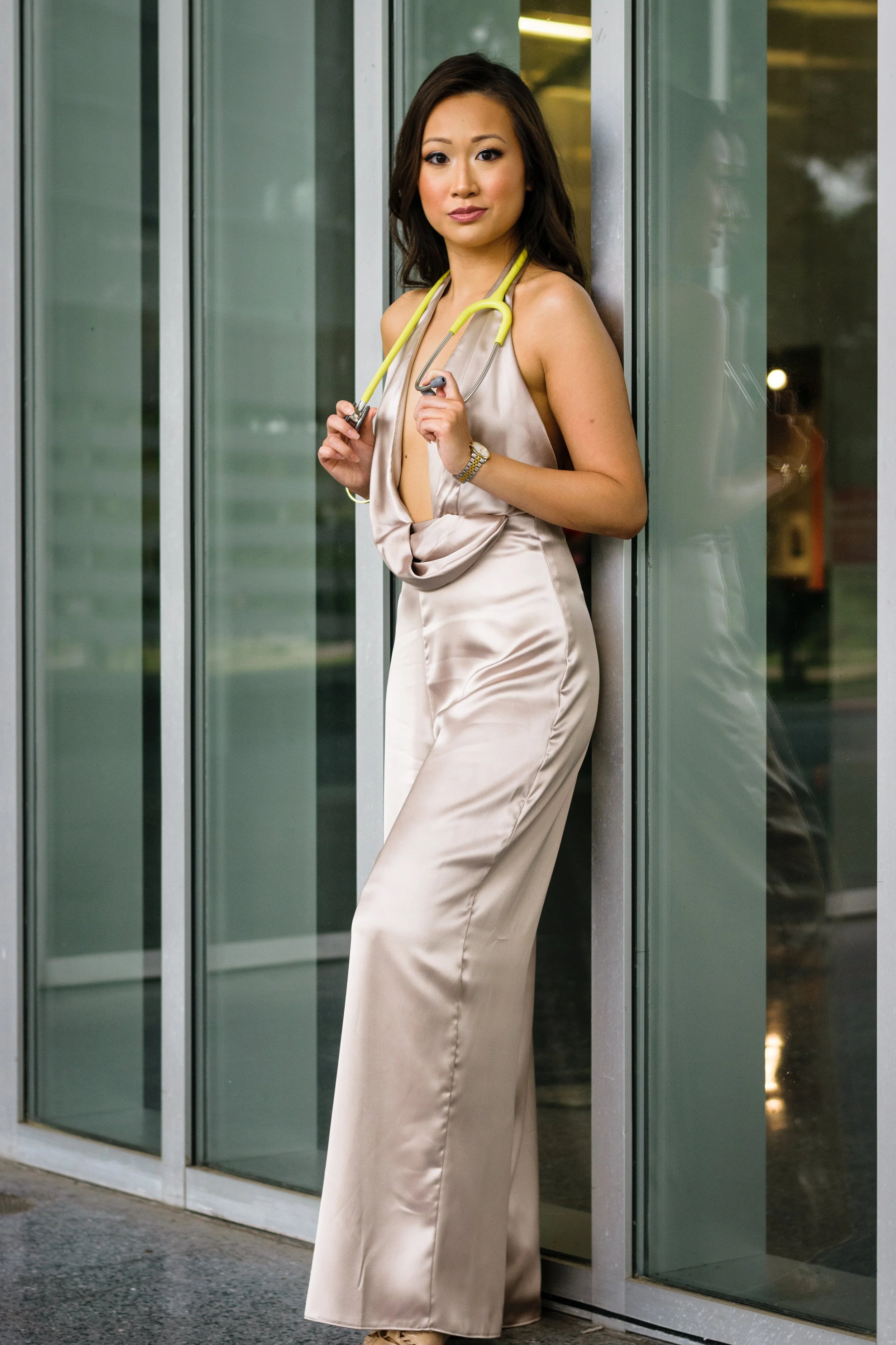 A woman in a silky beige dress holding a yellow stethoscope, standing outside near glass doors, with her reflection visible.